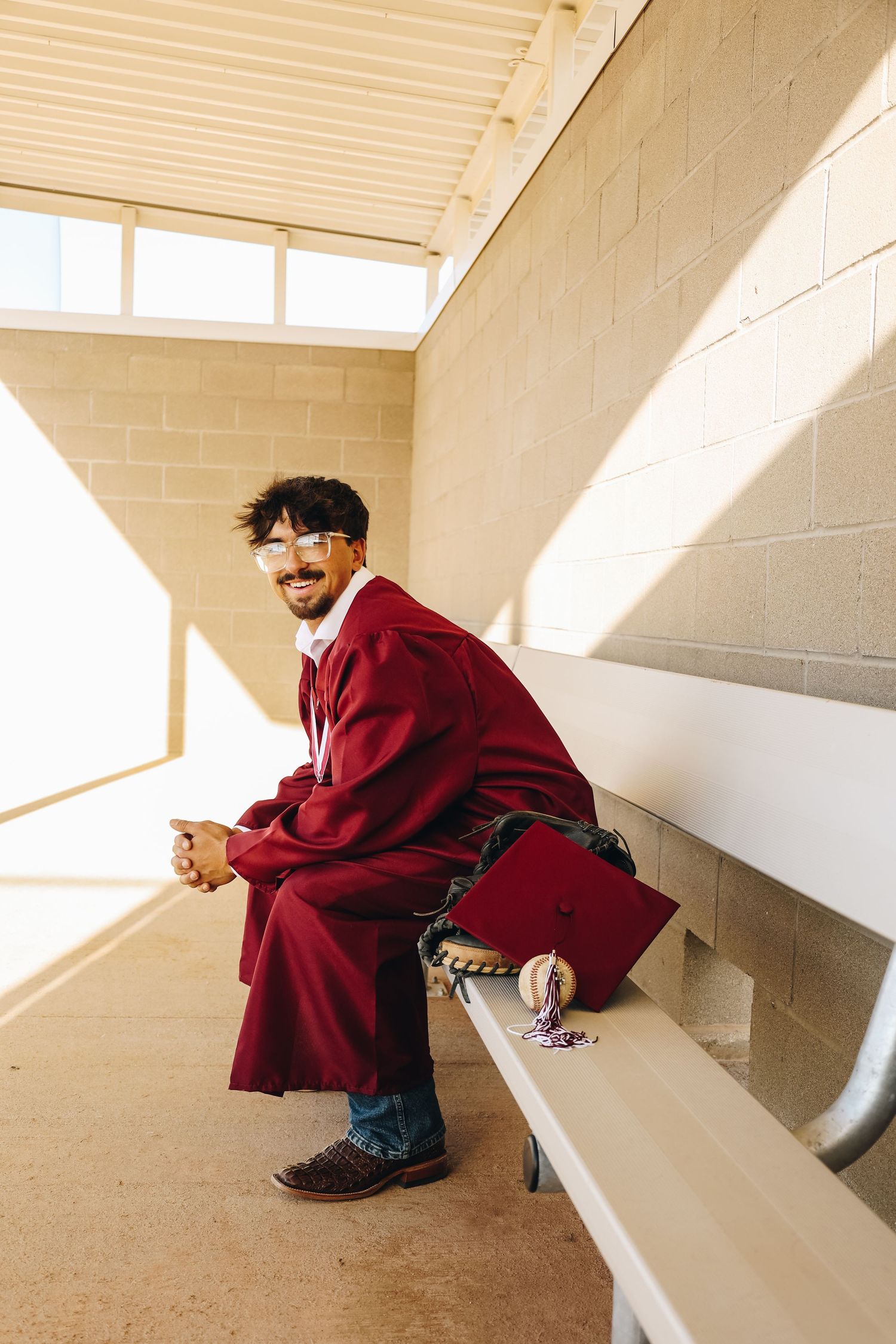 Graduate in maroon cap and gown sitting on bleachers smiling in sunlit outdoor corridor.
