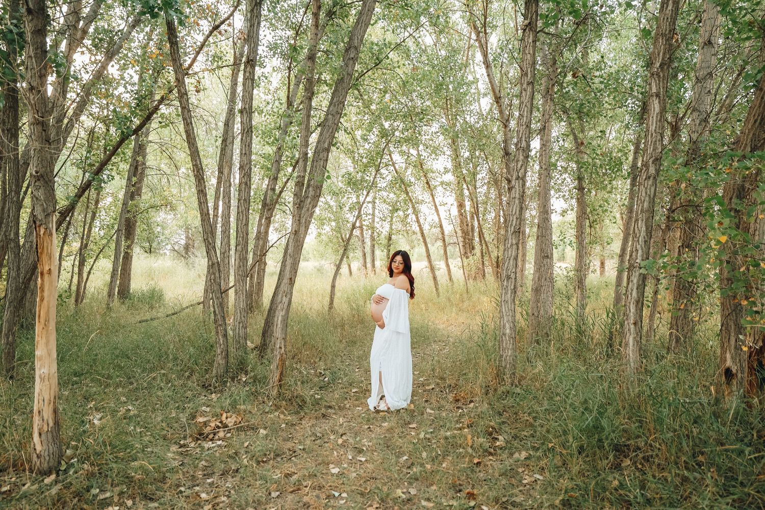 Pregnant woman in white maxi dress standing on forest path surrounded by tall slender trees.