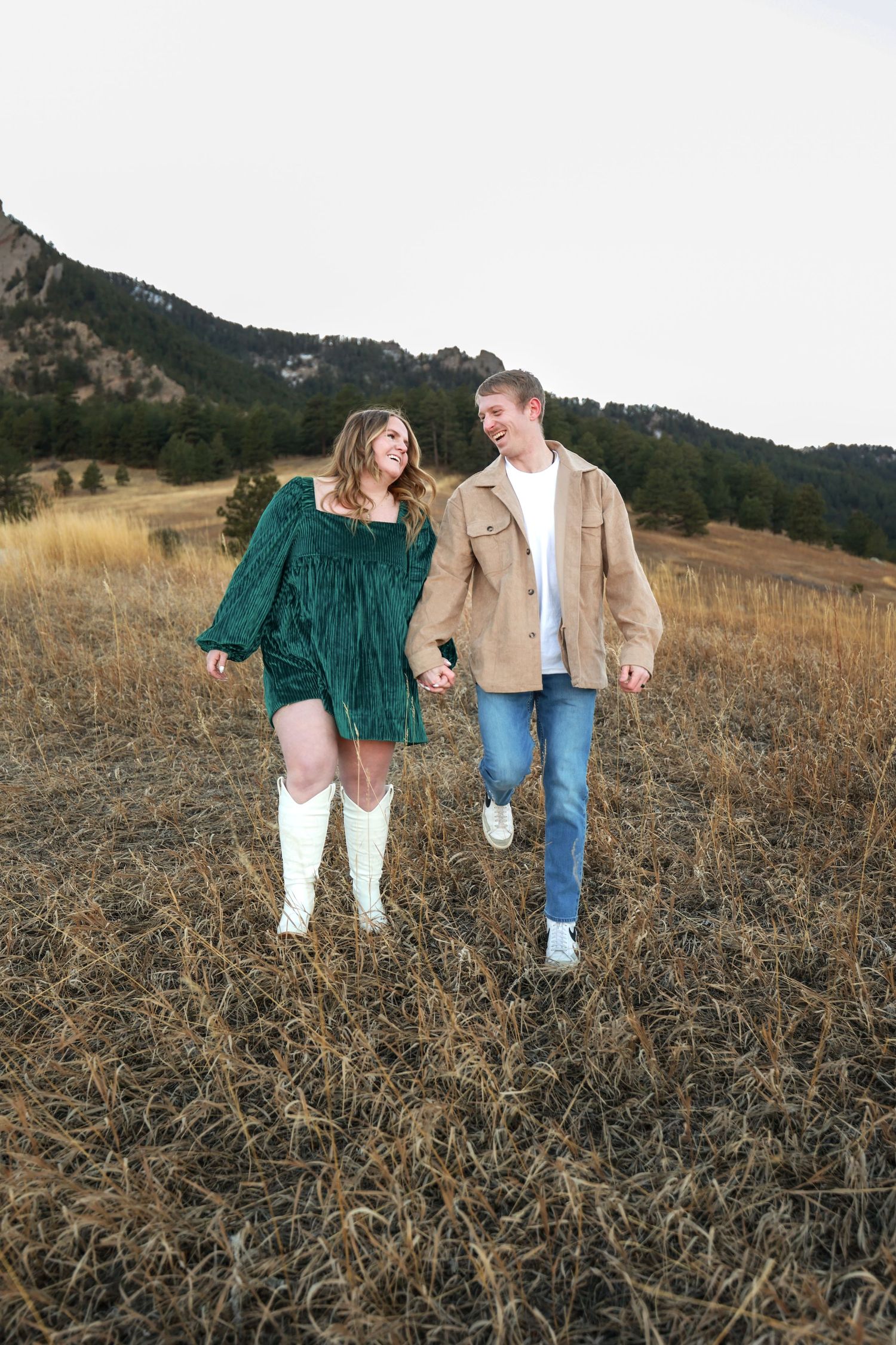 Couple walking hand in hand through dry grass field, woman in green dress and white boots with mountain backdrop.