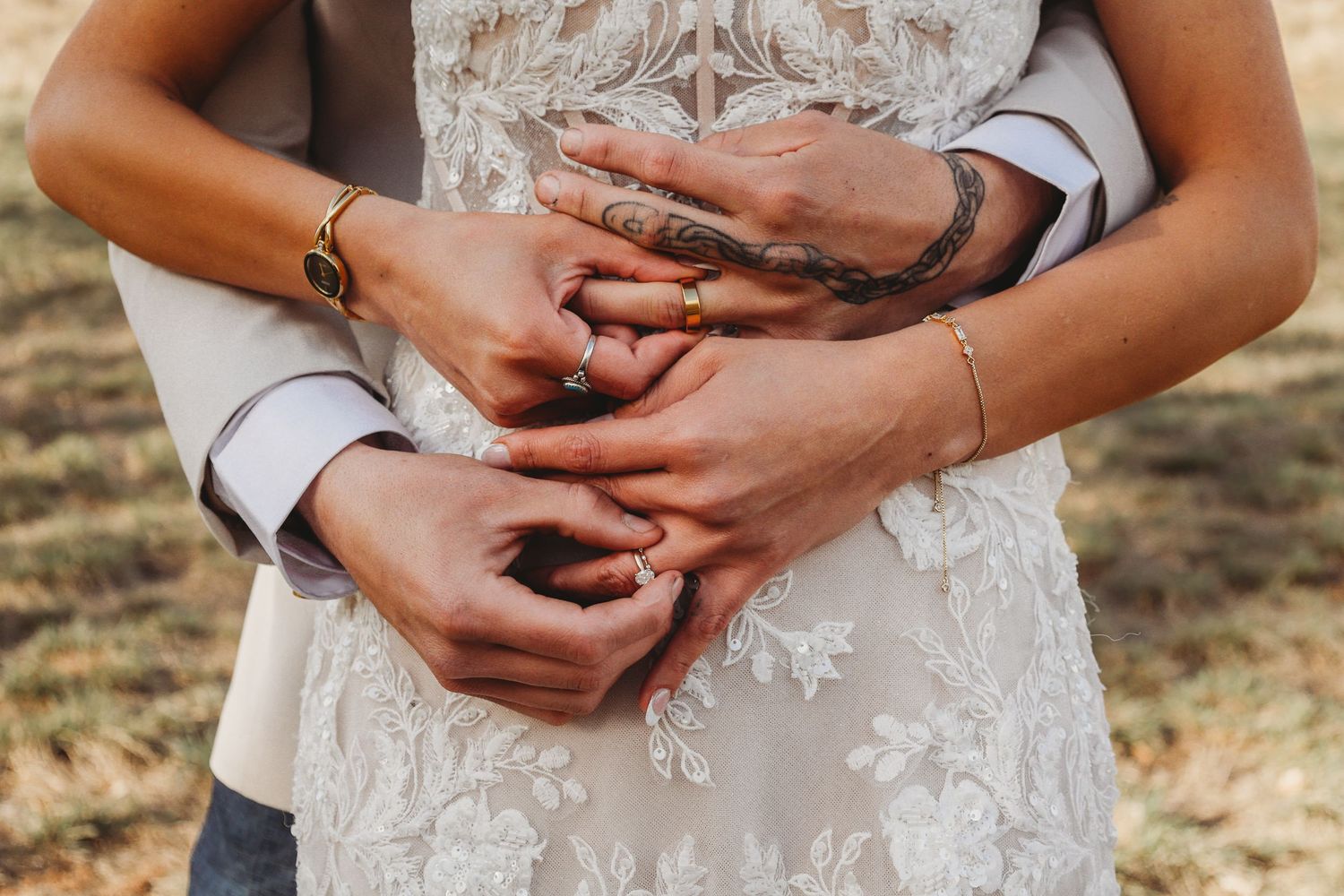 Close-up of bride and groom holding hands together against her lace wedding gown, showing wedding rings.