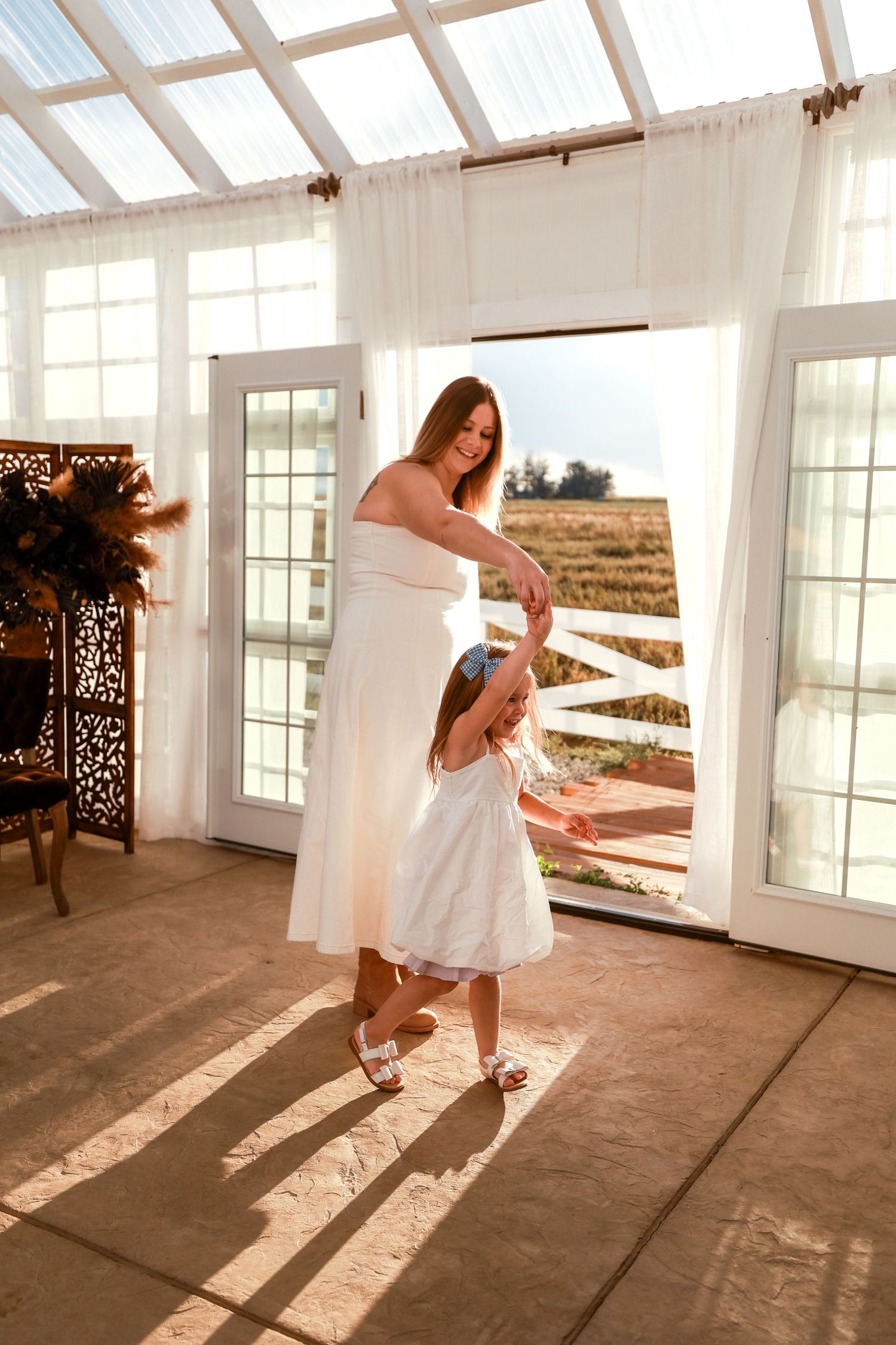 Mother in white dress spinning young daughter in white dress inside a sunlit greenhouse.