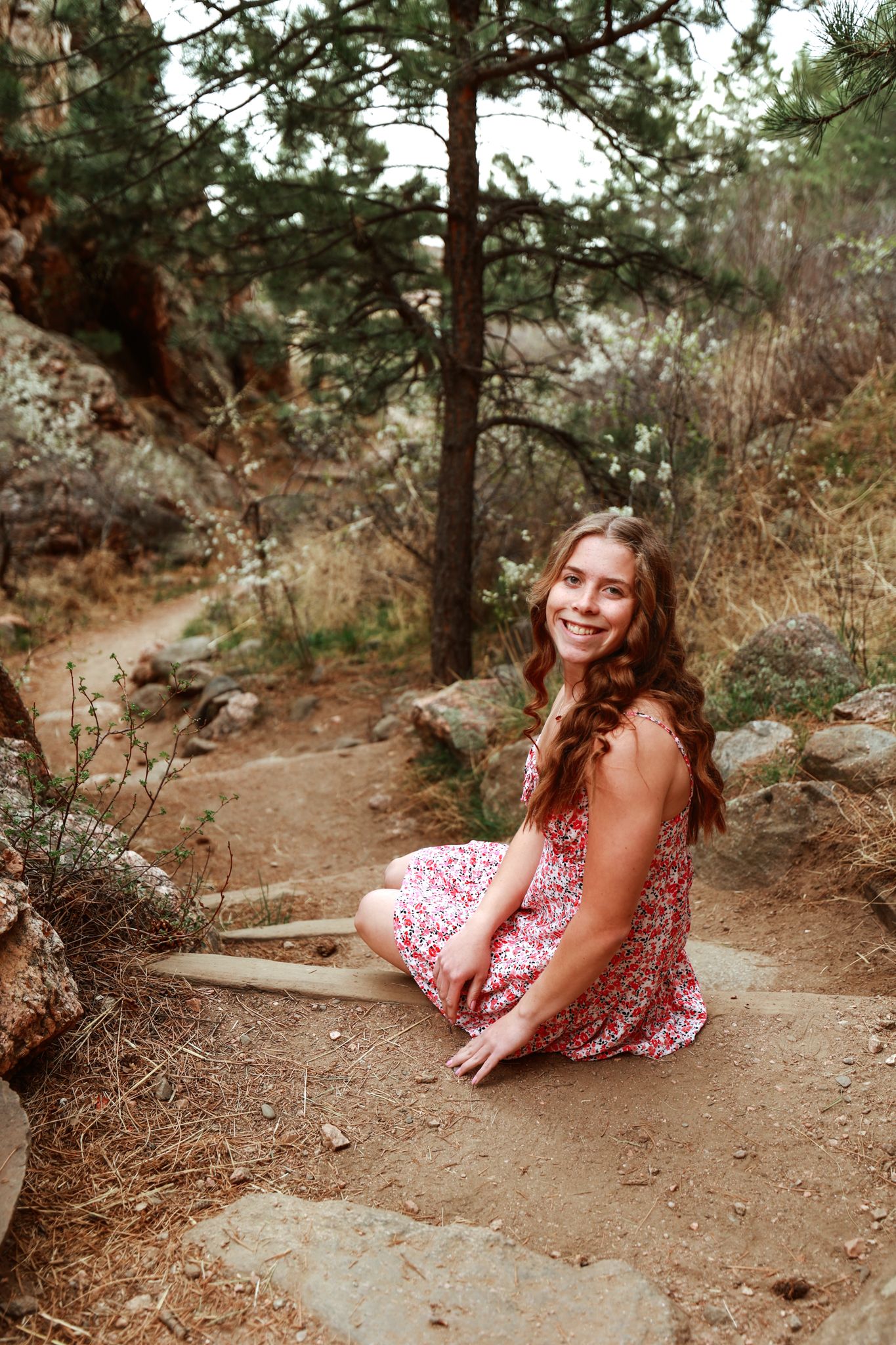 Young woman in floral dress sitting on rocky trail, smiling in a pine forest setting.