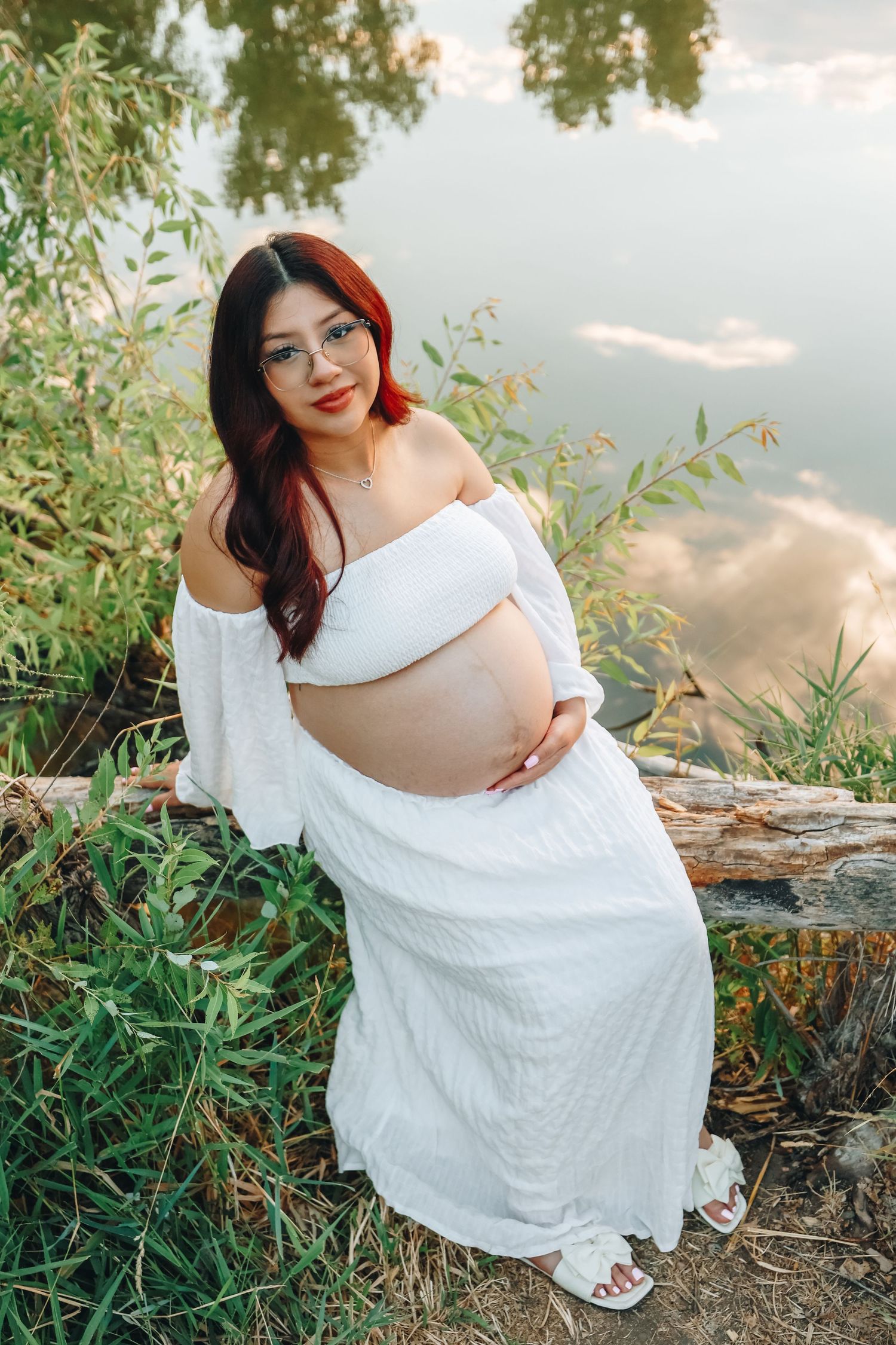 Smiling pregnant woman with red hair in white dress sitting on log near lakeside greenery.