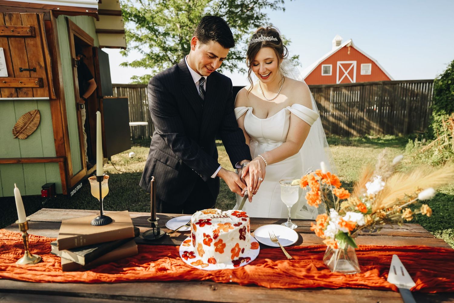 Bride and groom smiling while cutting a floral-decorated wedding cake at a rustic outdoor table near a red barn.