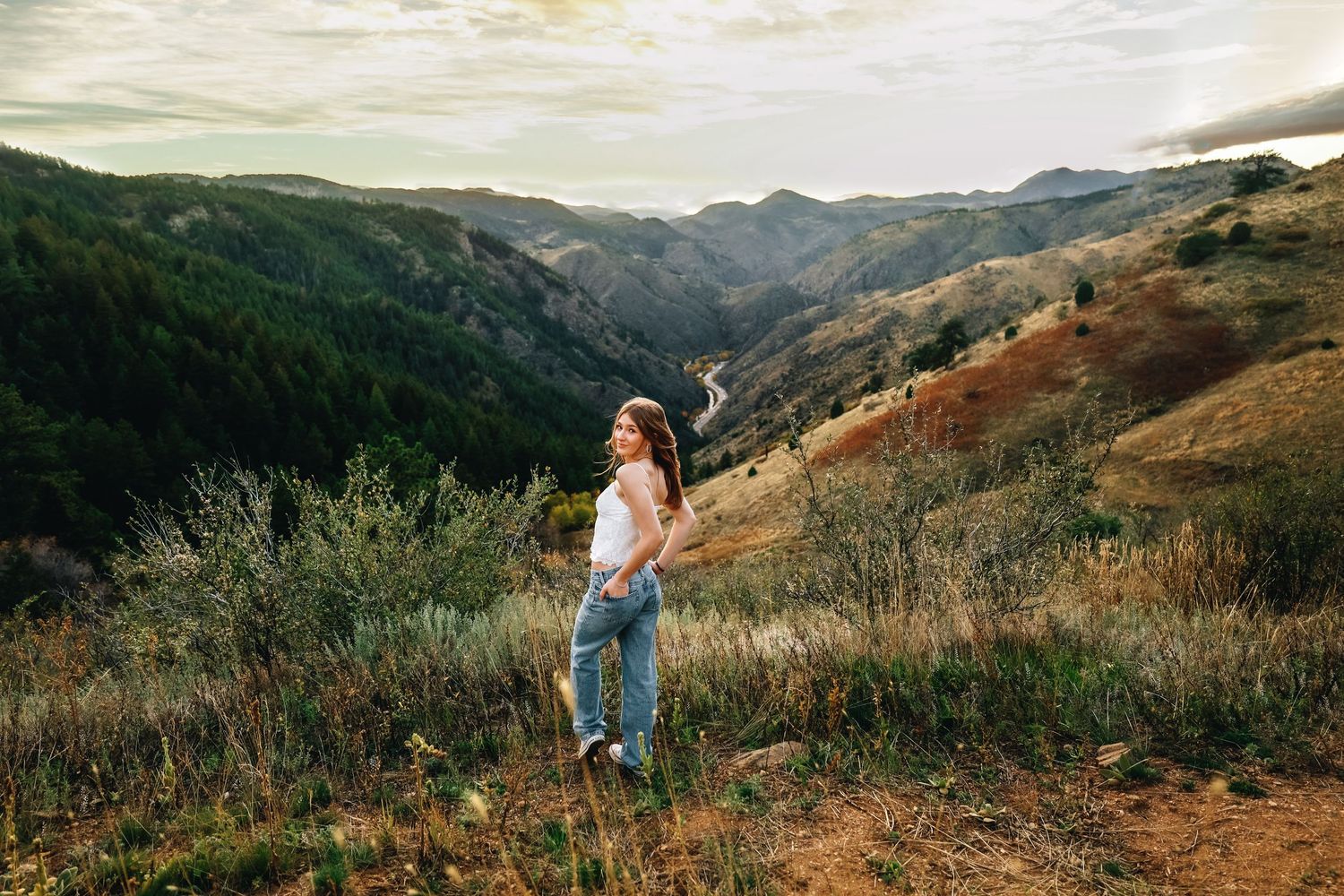 Young woman in white top and jeans standing on hillside overlooking scenic mountain valley.