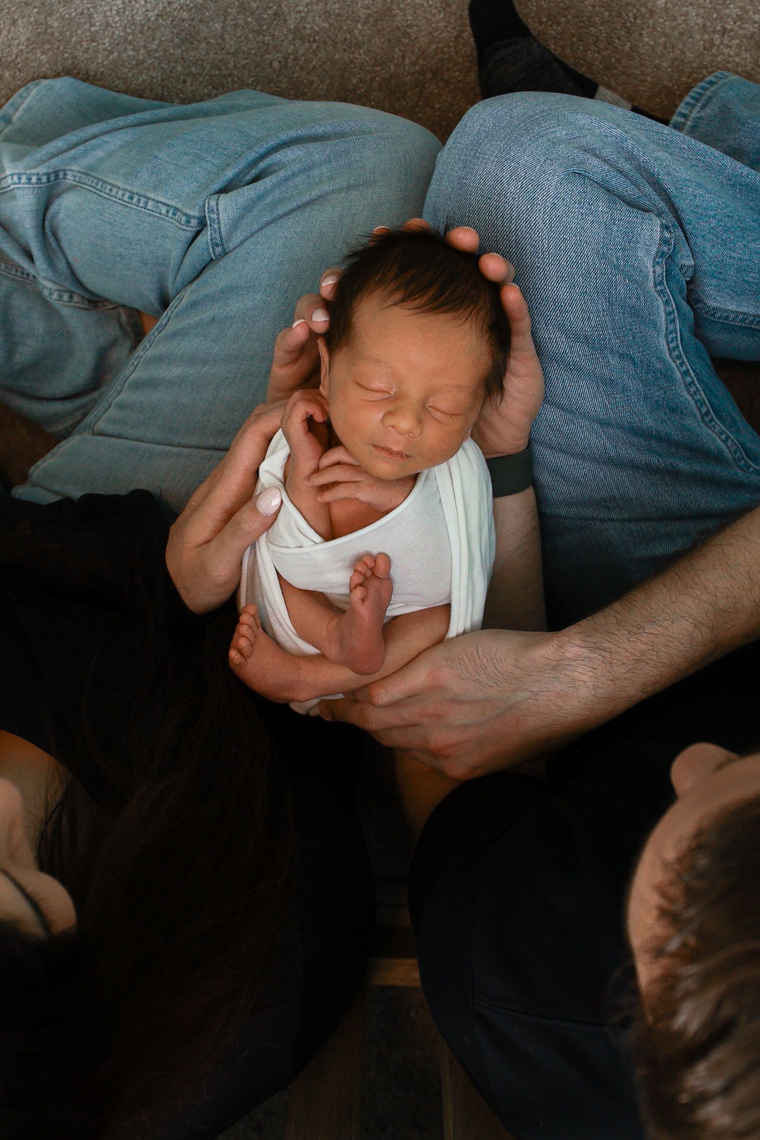 Sleeping newborn in white swaddle cradled gently between two adults wearing jeans.