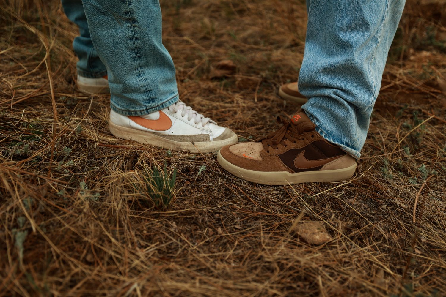 Two people wearing Nike sneakers standing on pine needle covered ground outdoors.