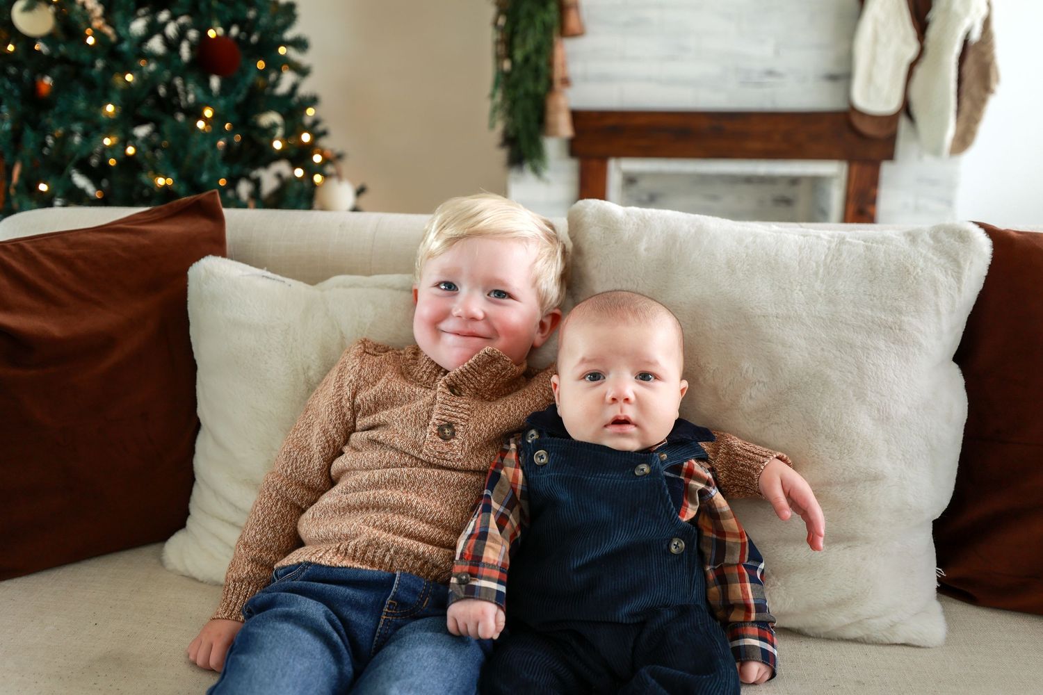 Two young brothers pose on a cream sofa with Christmas tree and festive fireplace mantel in background.