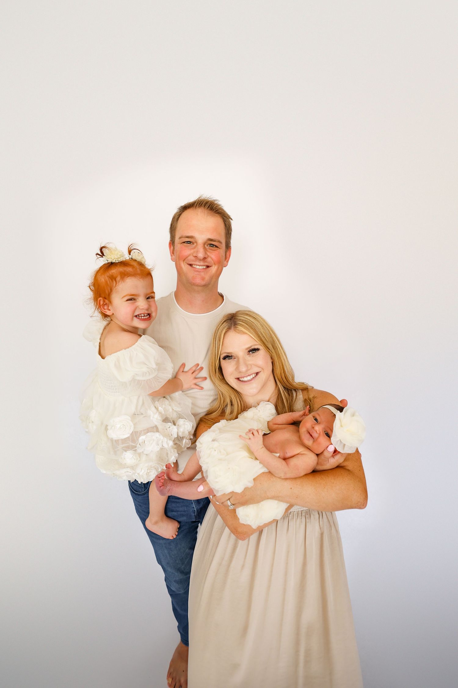 Happy family portrait with father, mother holding newborn, and red-haired toddler daughter in white dress.