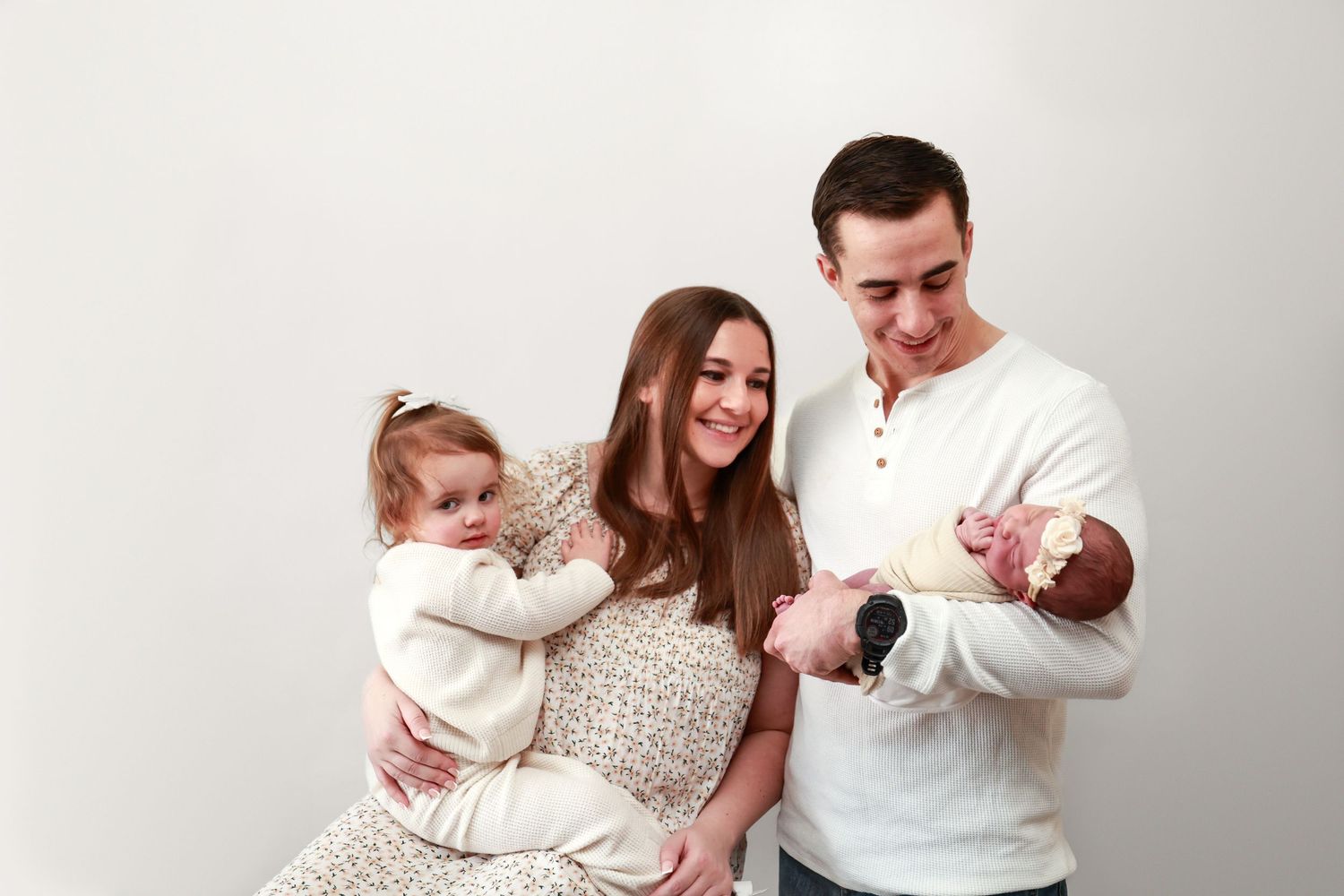 Happy young family with mother holding toddler girl and father cradling newborn baby, posing together against white background.