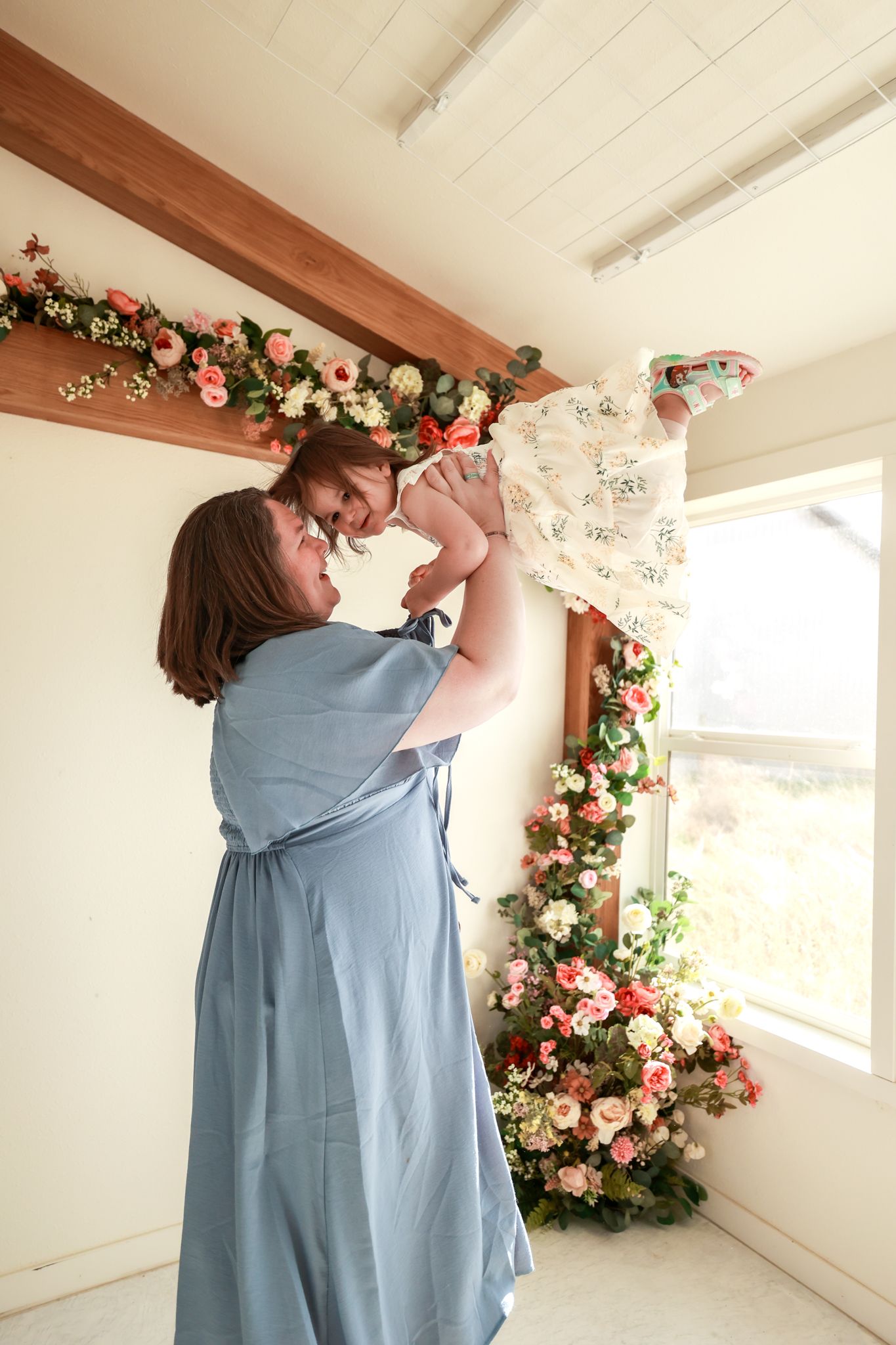 Mother in blue dress lifts smiling baby in white lace dress amid floral decorations in bright studio.