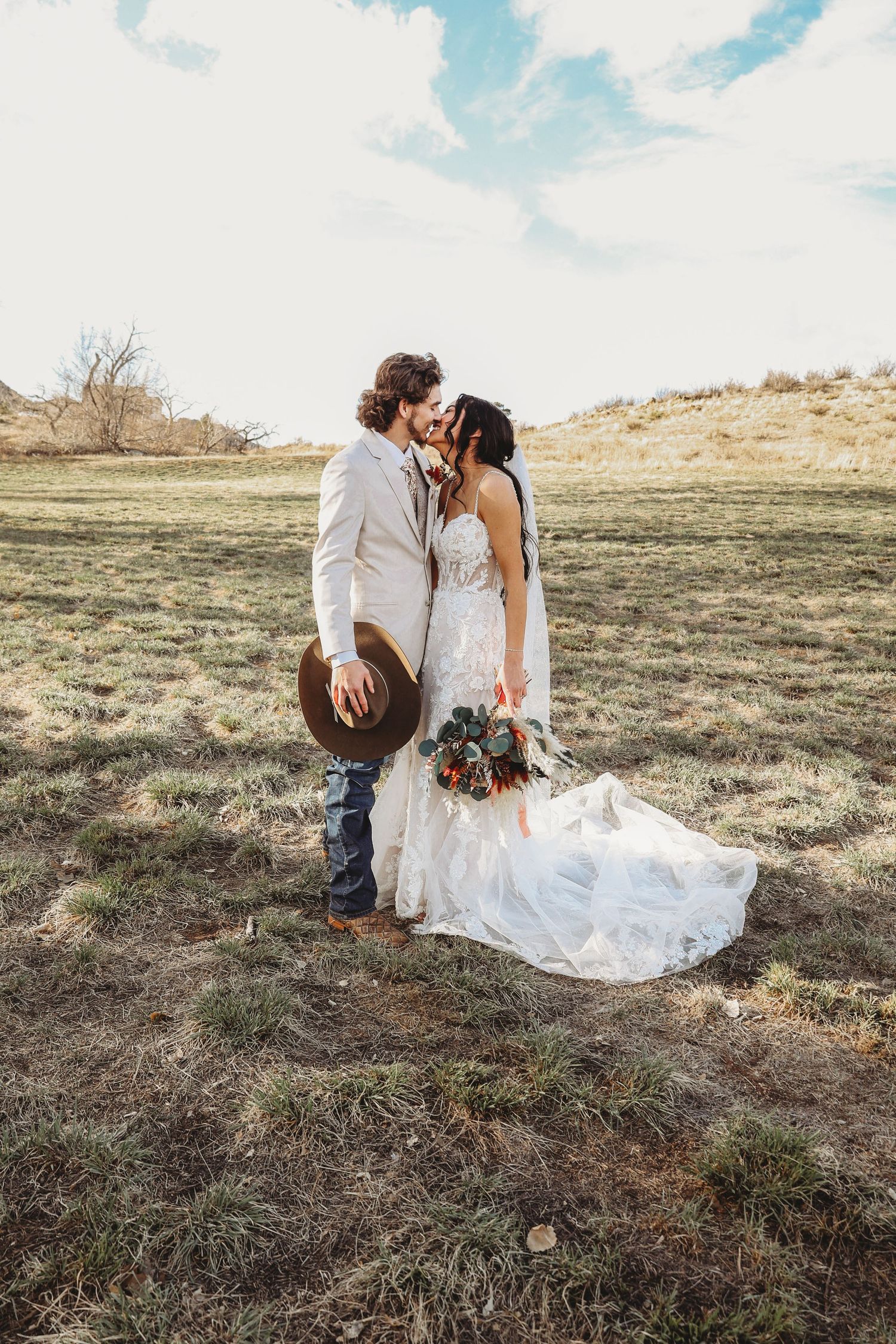 Bride and groom kissing in a rustic open field, groom holding a brown cowboy hat and bride in a flowing lace gown.