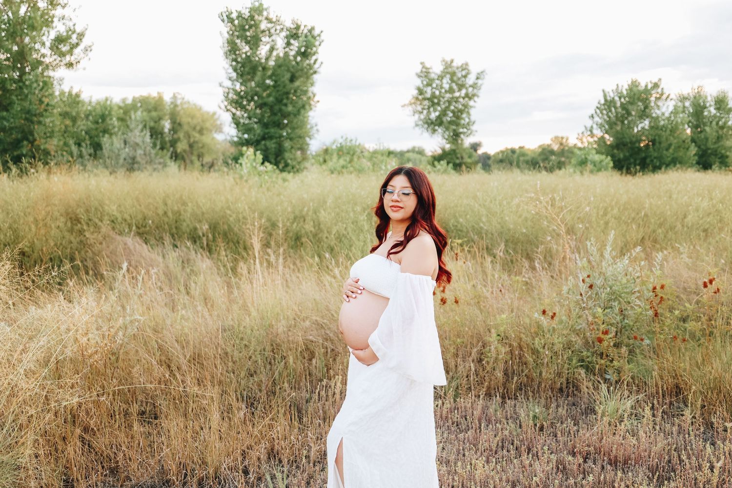 Smiling pregnant woman in white dress cradling baby bump while standing in a grassy meadow.