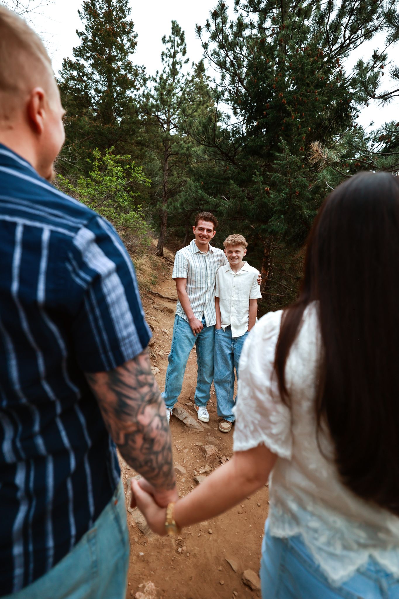 Family of four on a forest trail, parents holding hands facing two smiling boys among pine trees.