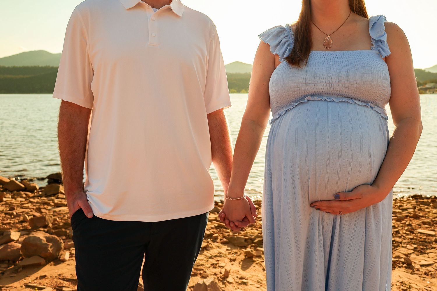 Couple holding hands at sunset by a lake, pregnant woman in blue dress cradling baby bump.