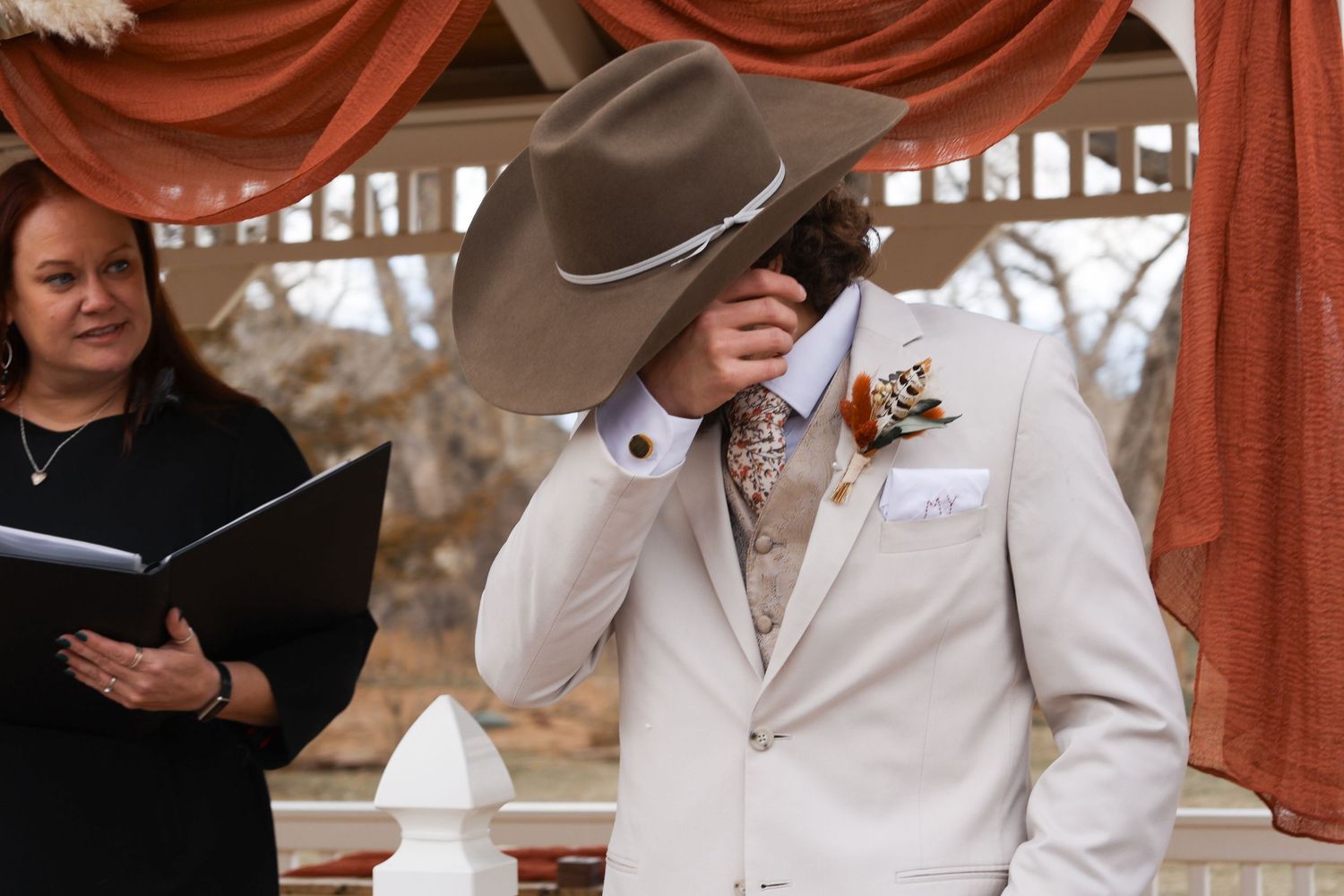 Emotional groom hiding face under cowboy hat during outdoor wedding ceremony.