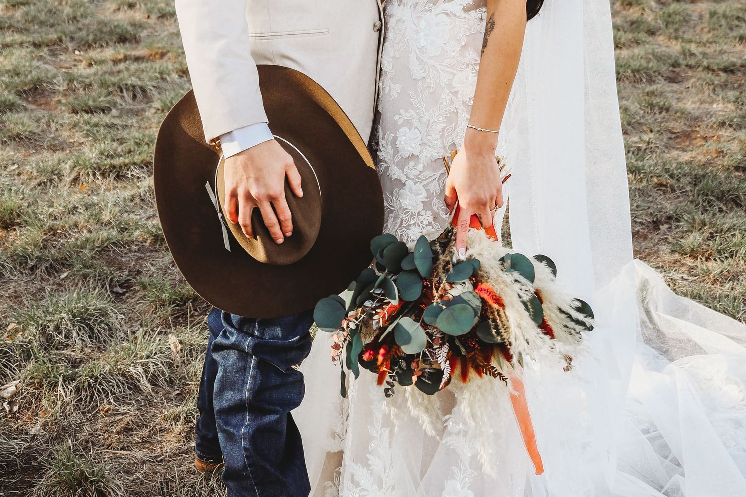 Bride and groom holding cowboy hat and floral bouquet with eucalyptus in a field.