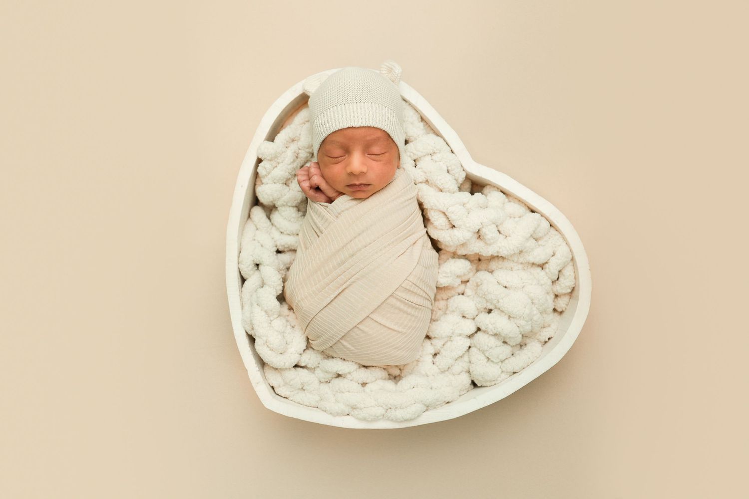 Newborn baby swaddled in cream wrap with knot hat sleeping in white heart-shaped bowl with chunky knit blanket.