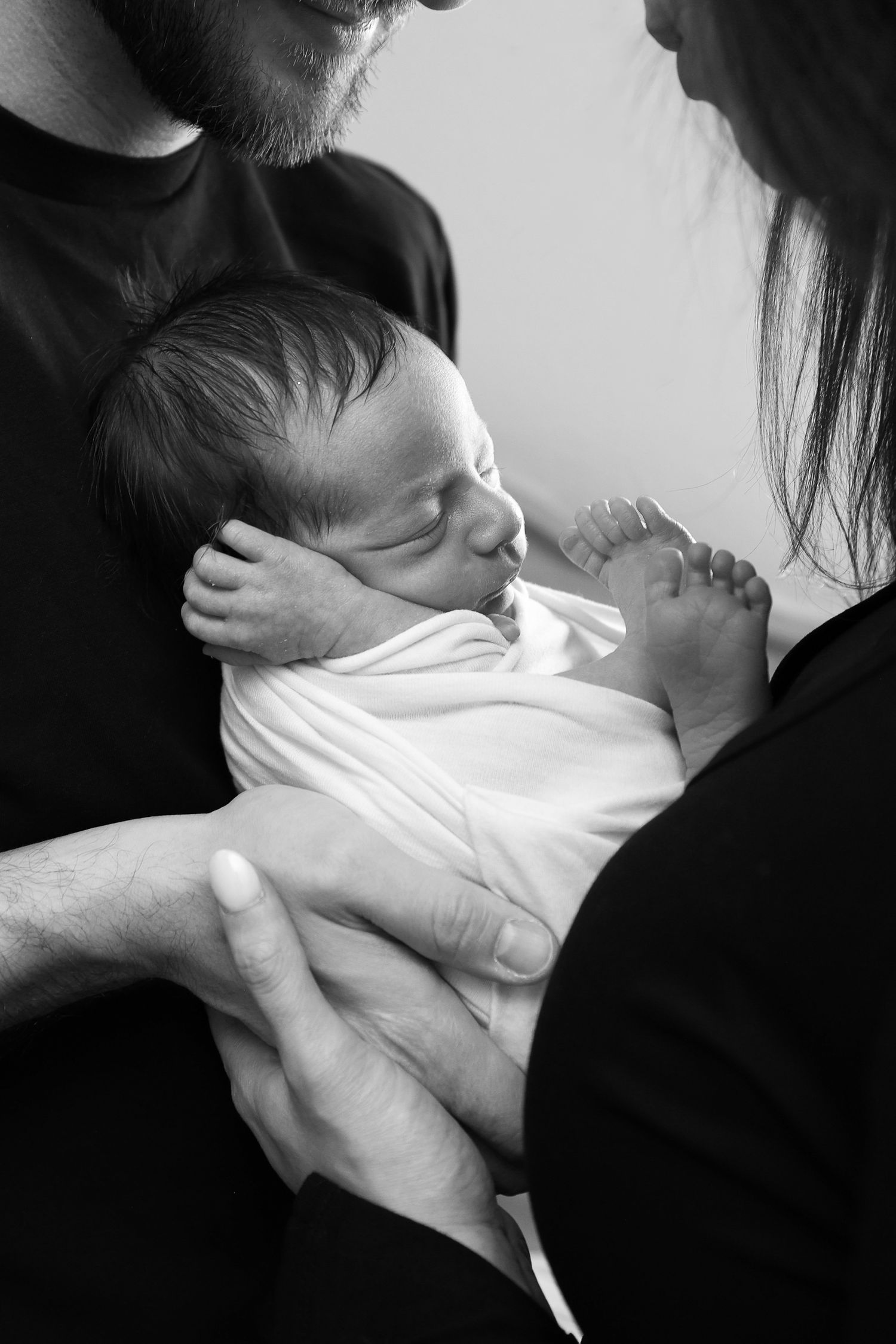 Black and white close-up of parents holding swaddled sleeping newborn baby together.