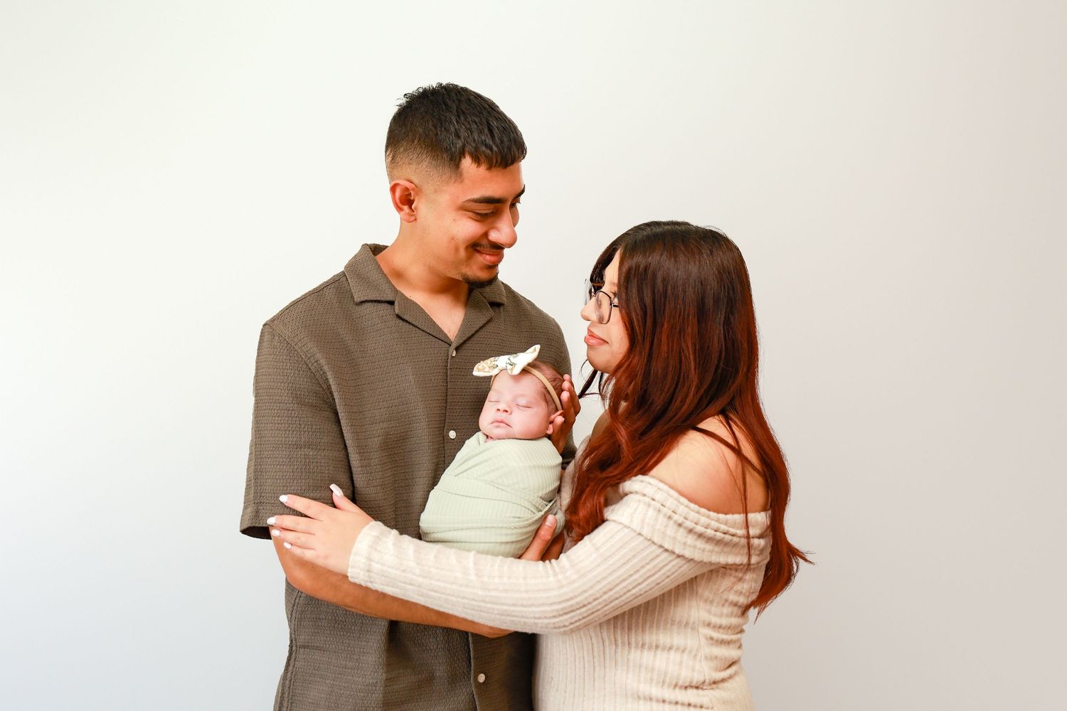 A couple smiles at their newborn baby wrapped in green, posed against a white background.