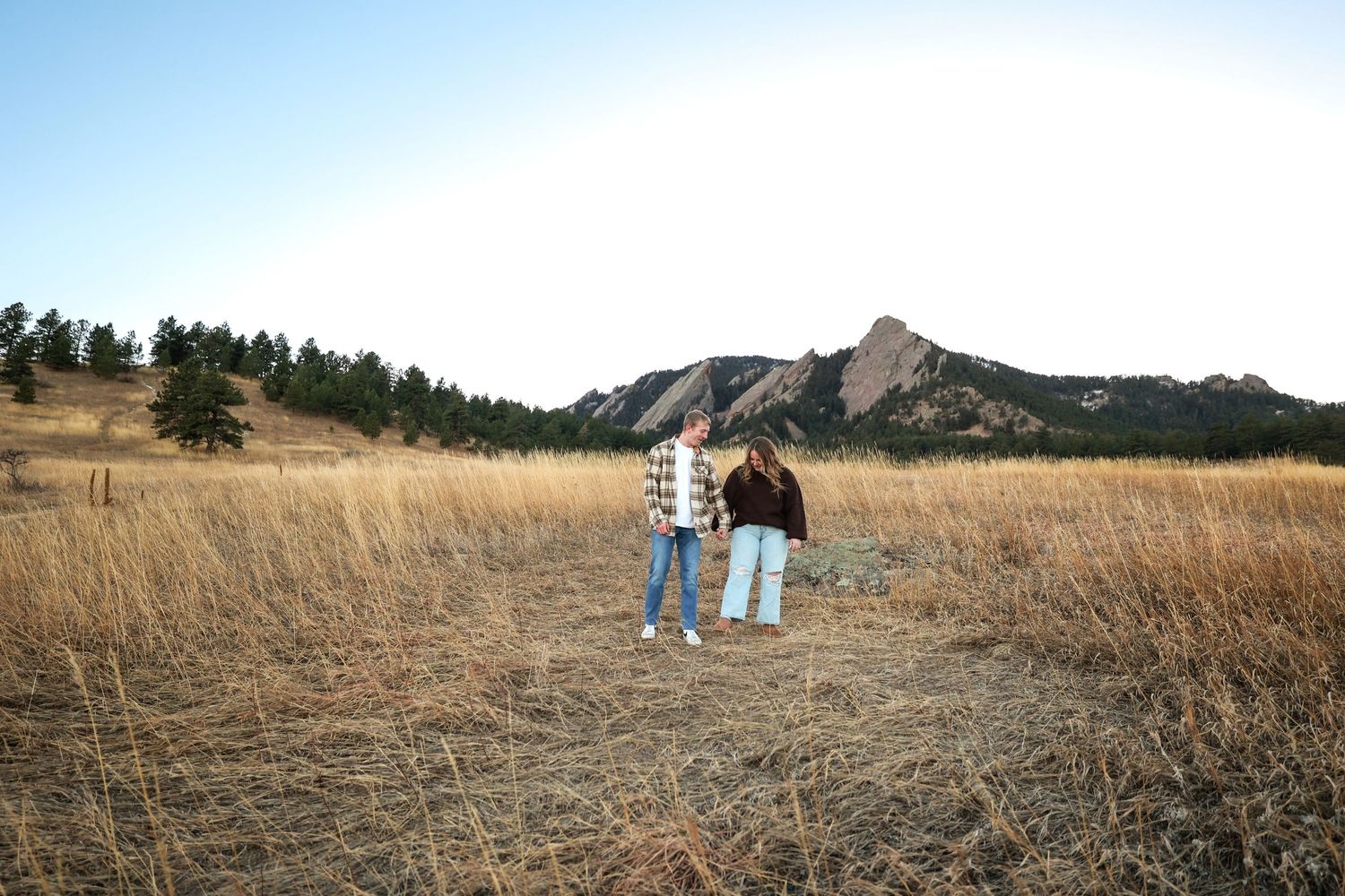 Couple standing together in wide open golden field with iconic Colorado Flatirons rock formations in background.