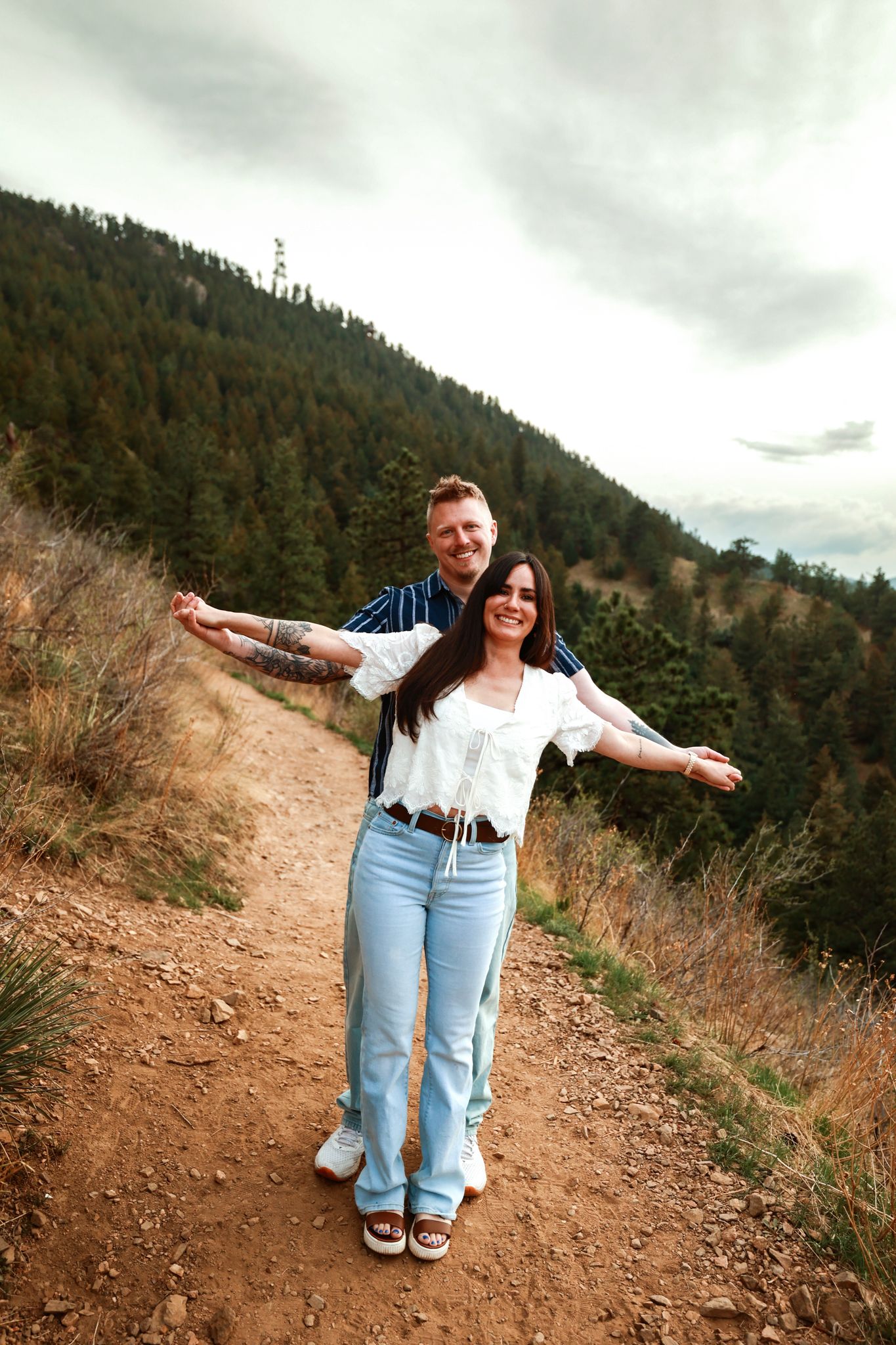 A joyful couple poses on a mountain hiking trail, arms outstretched, surrounded by pine trees and overcast skies.