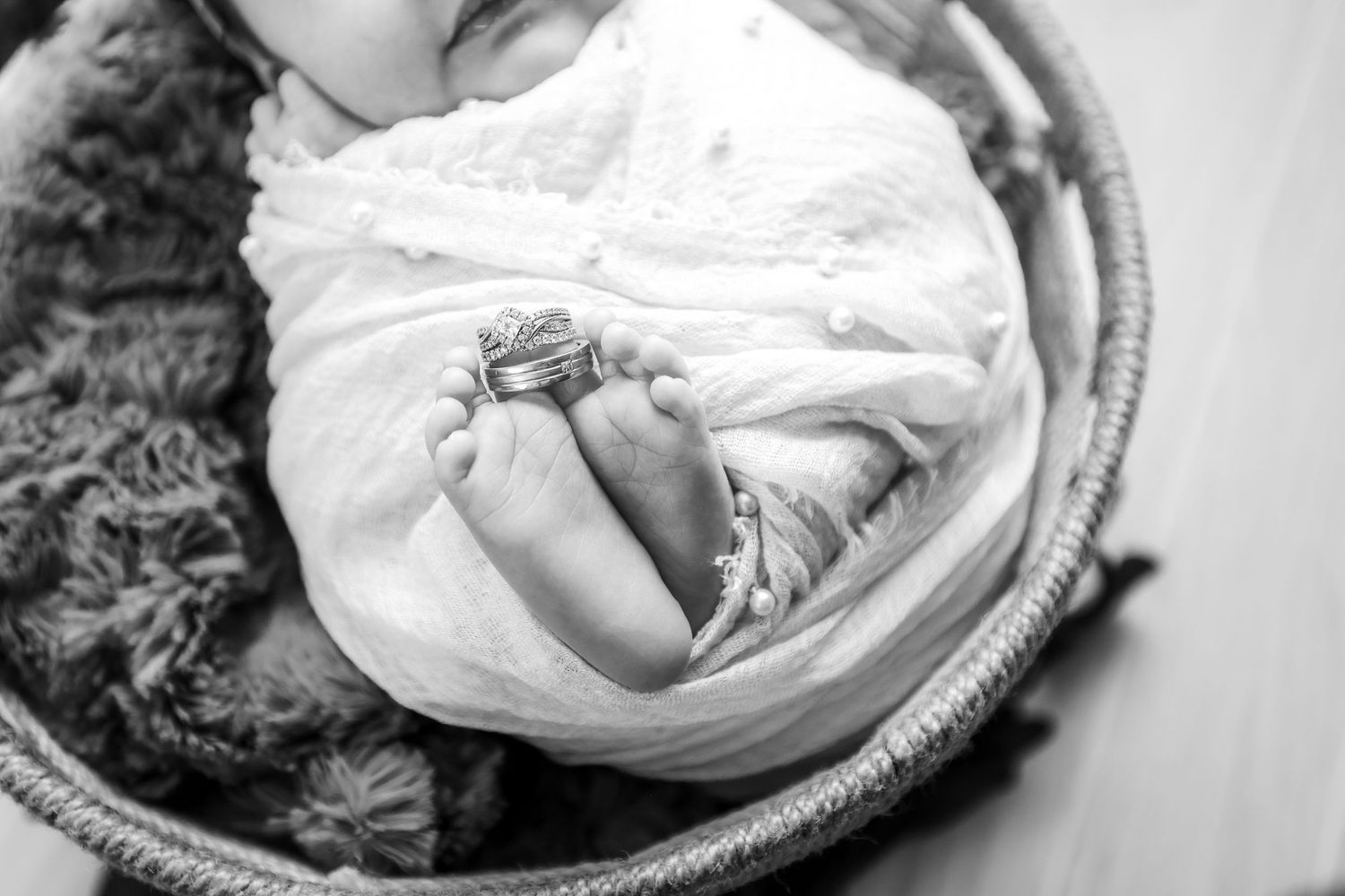 Black and white newborn photo of tiny baby feet wrapped in white cloth with rings placed on toes in a wicker basket.