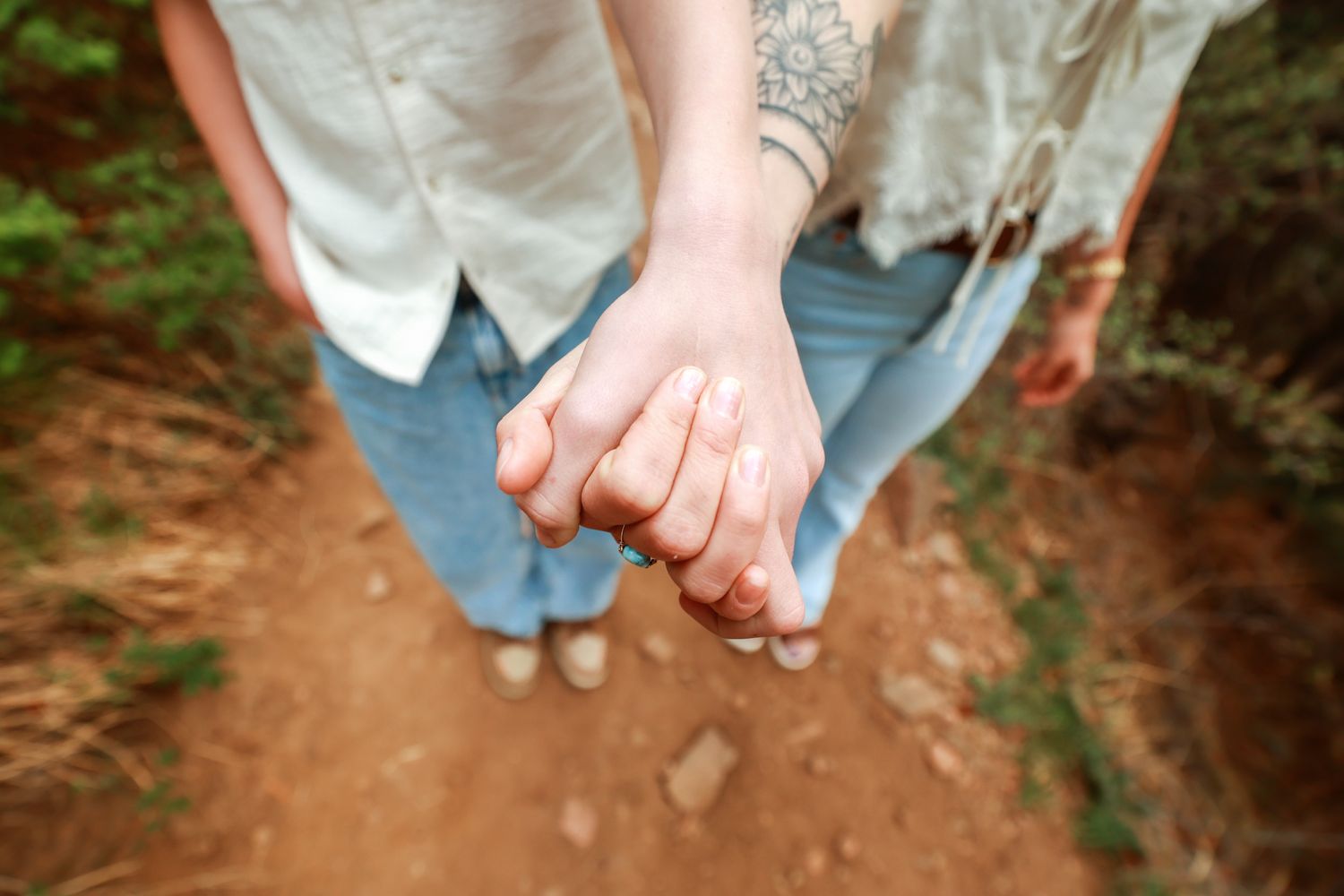 Two people holding hands on a dirt trail, one with a floral tattoo on their wrist, wearing casual jeans.