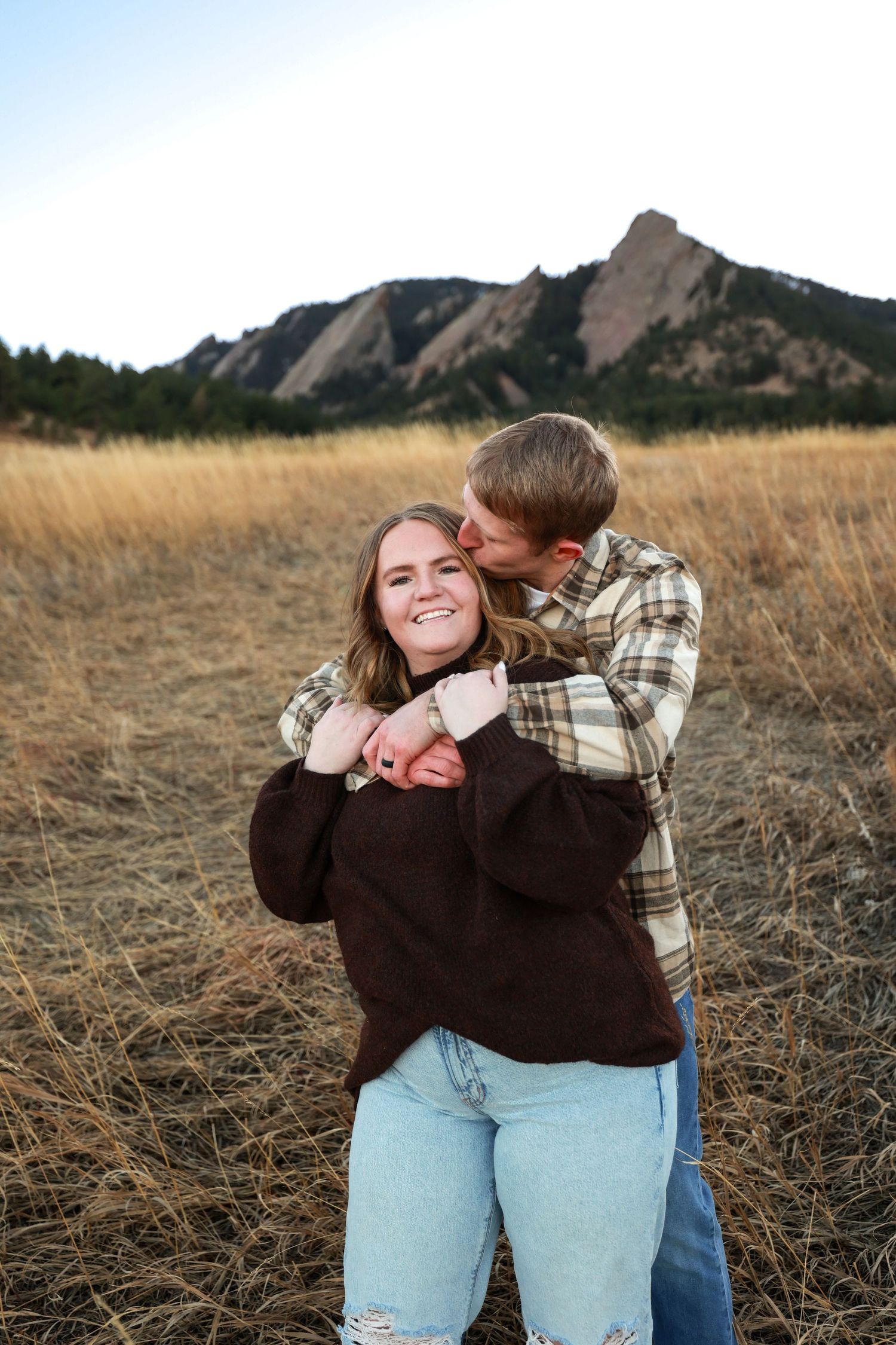Couple embracing in golden field with Colorado Flatirons mountain backdrop during autumn engagement photo session.