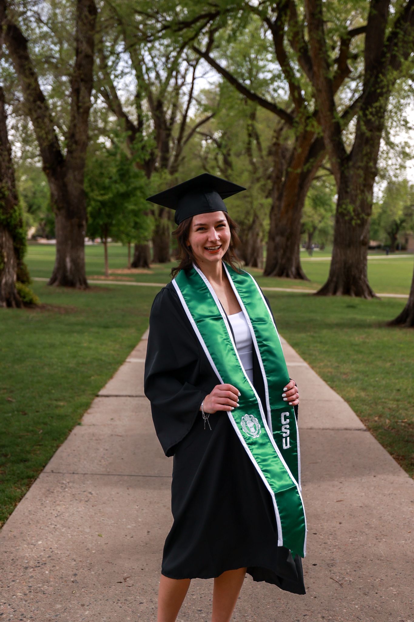 Smiling female graduate in black cap and gown with green sash poses on campus pathway surrounded by trees.