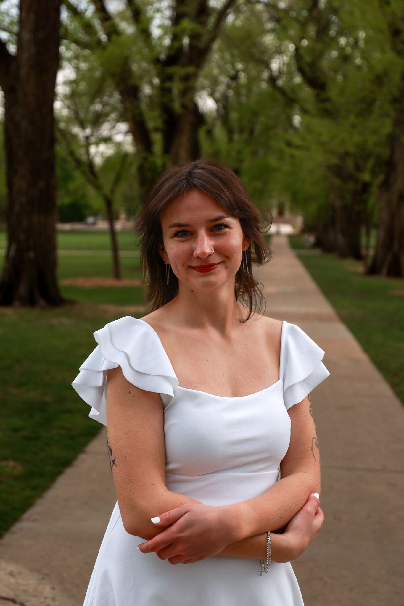 Young woman in white ruffle dress with tattoo stands smiling on tree-lined park pathway.