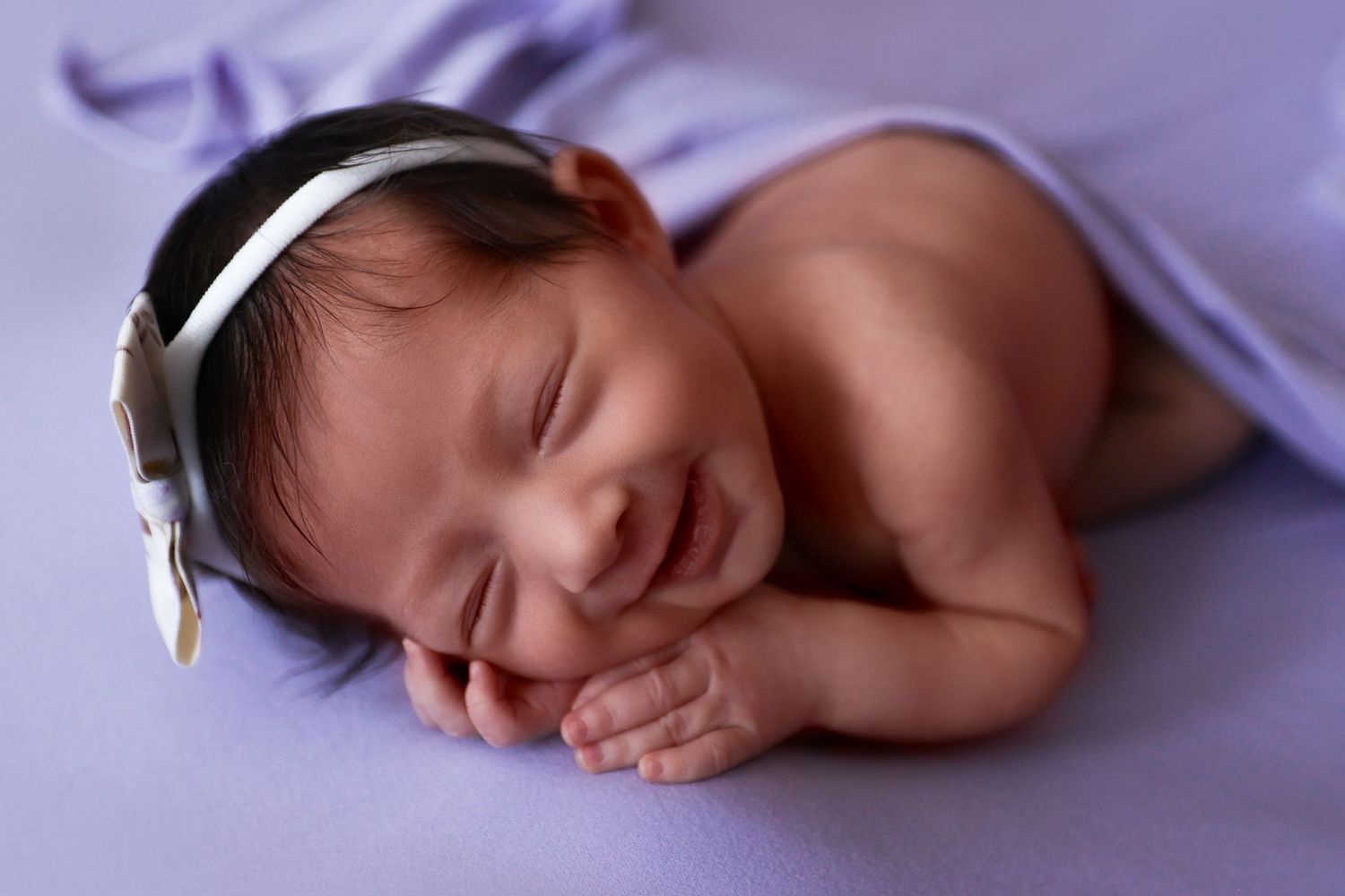 Adorable smiling newborn baby with white bow headband sleeping on a lavender blanket.