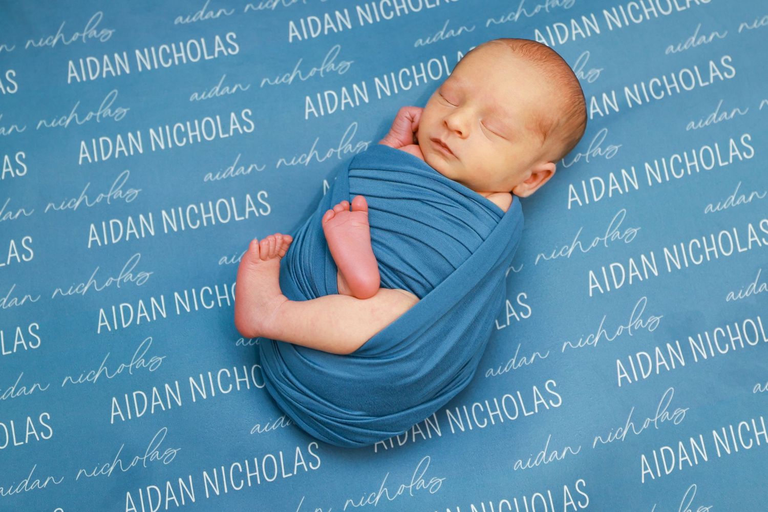 Newborn Aidan Nicholas wrapped in blue swaddle on a personalized name photo backdrop.