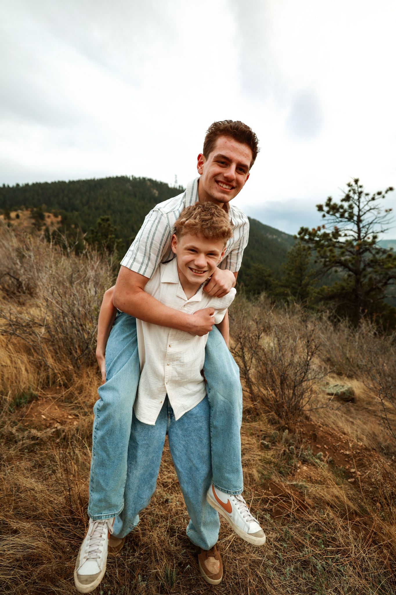 Two young men smiling outdoors on a mountain trail, one giving the other a piggyback ride.