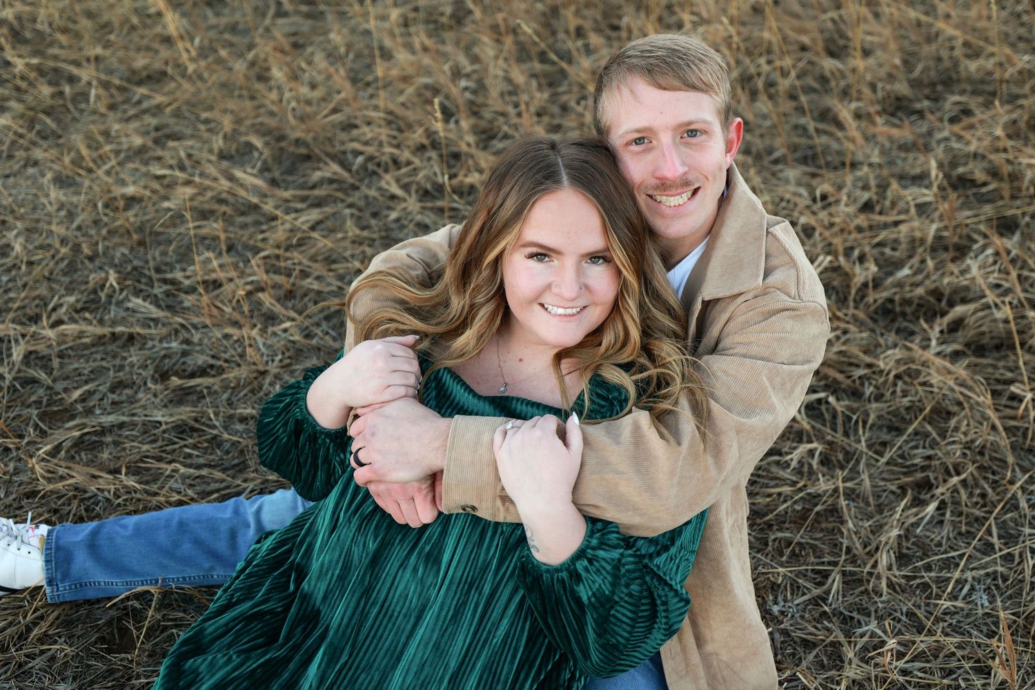 Couple posing together in dried grass field, woman in green velvet dress smiling during outdoor engagement session.