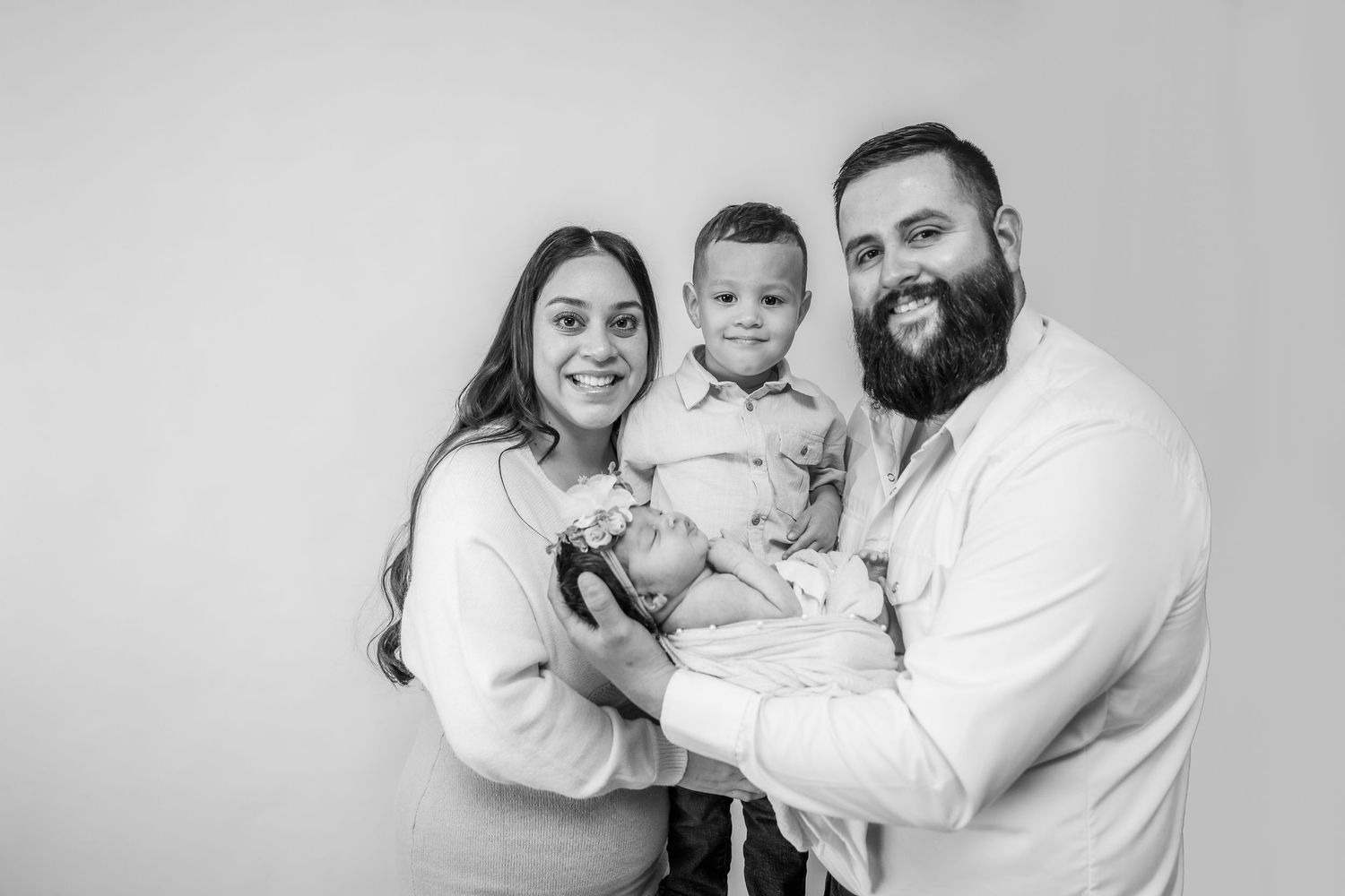 Black and white family portrait with parents holding newborn baby and toddler son, smiling warmly.