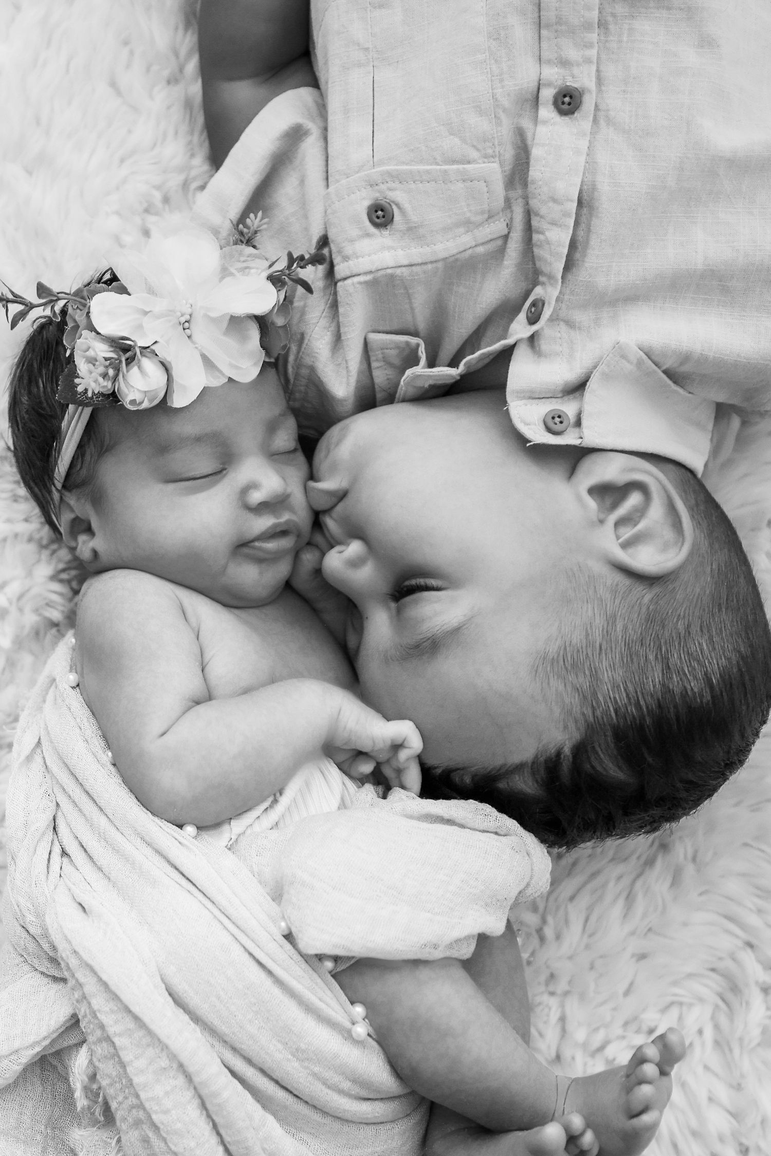Black and white photo of a young child gently kissing a sleeping newborn baby wearing a floral headband.