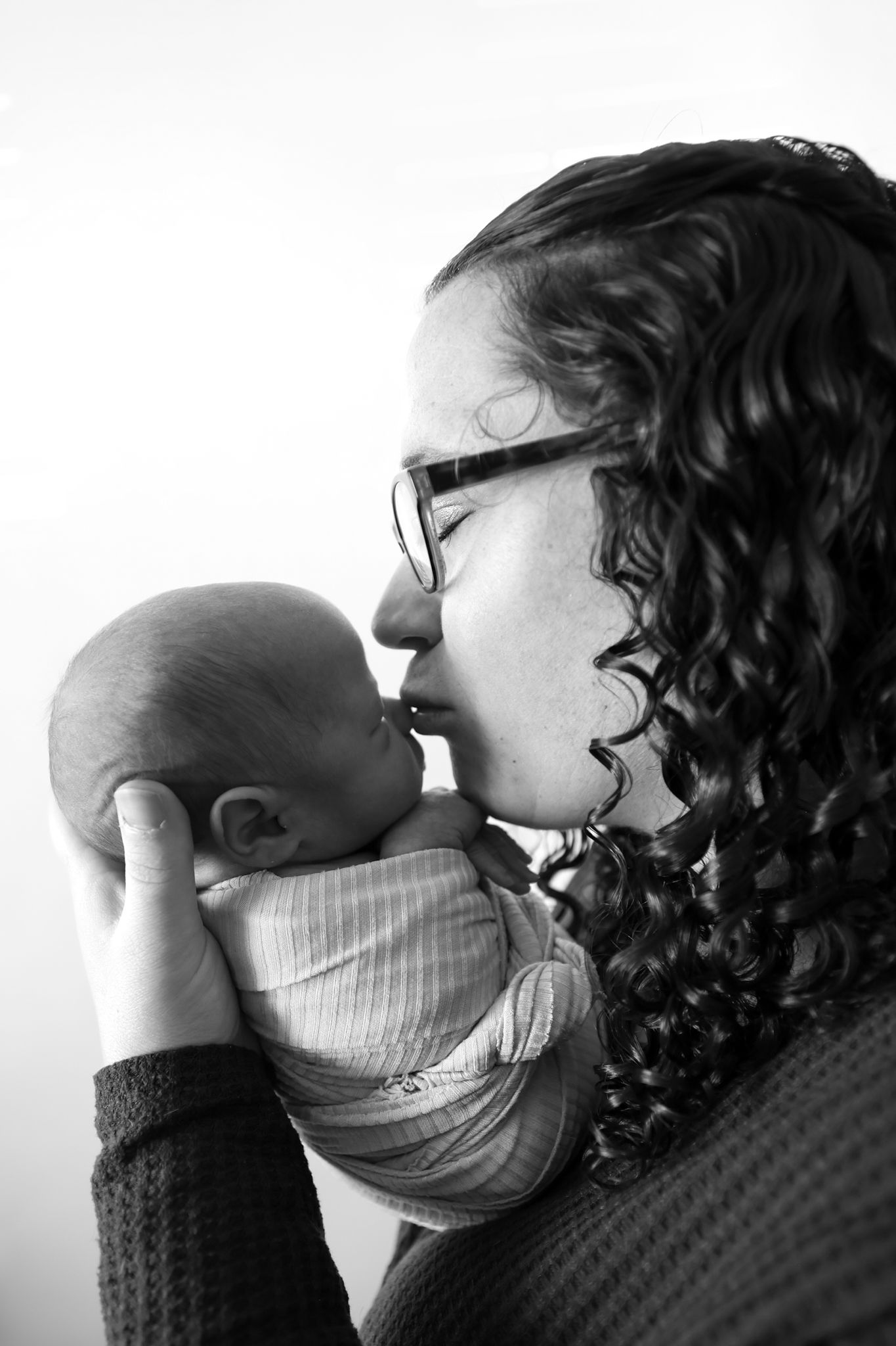Black and white photo of a curly-haired mother with glasses tenderly kissing her newborn baby.