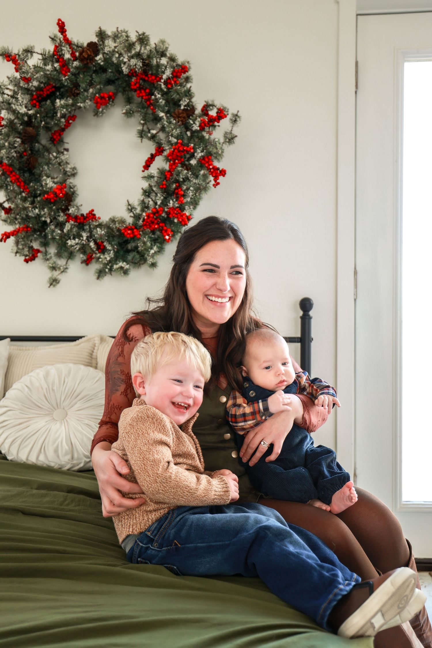 Smiling mother sitting on bed with two young sons near a festive Christmas wreath on the wall.