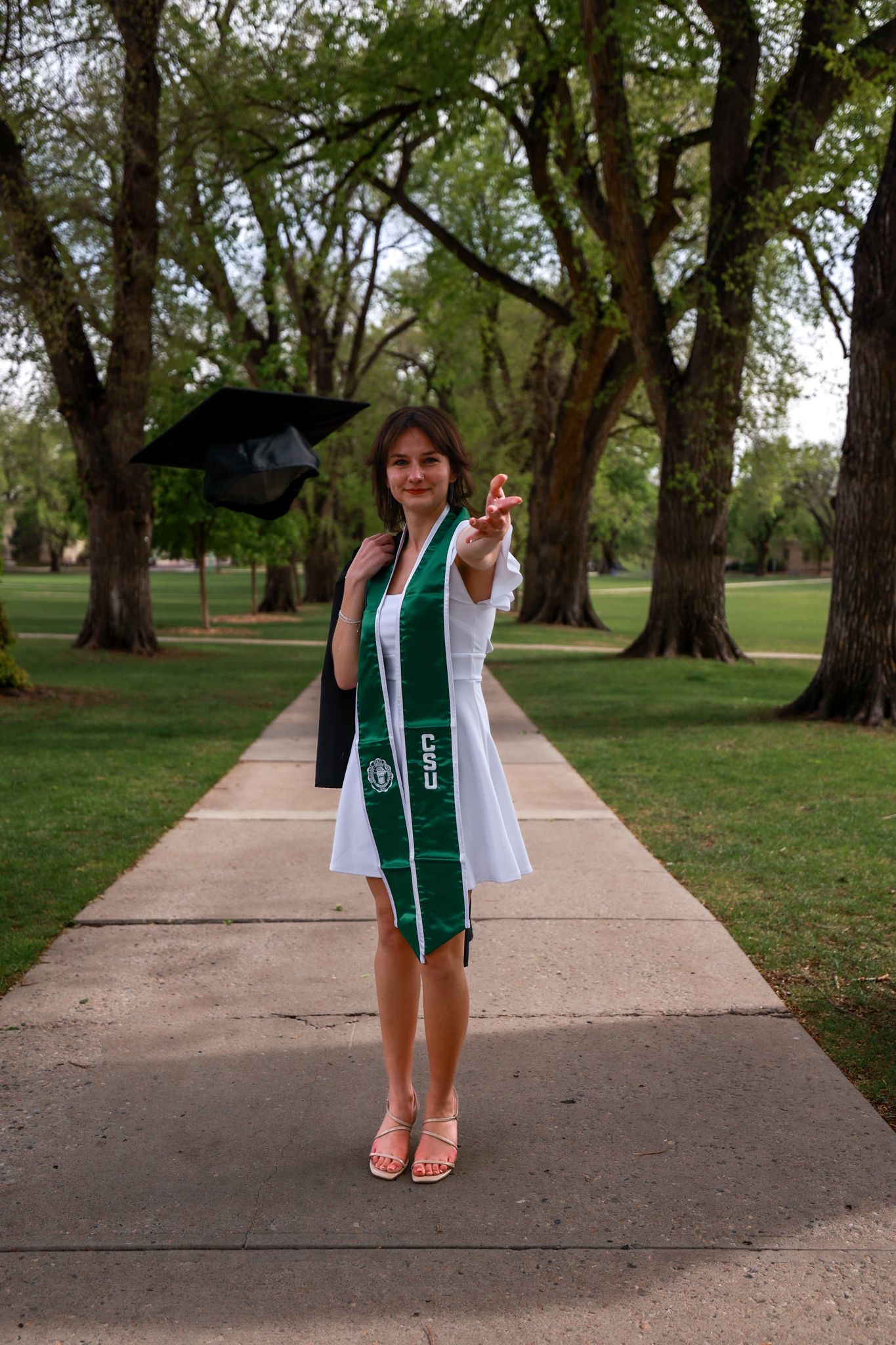 Young woman in white dress with green graduation stole holds cap outdoors on tree-lined campus walkway.