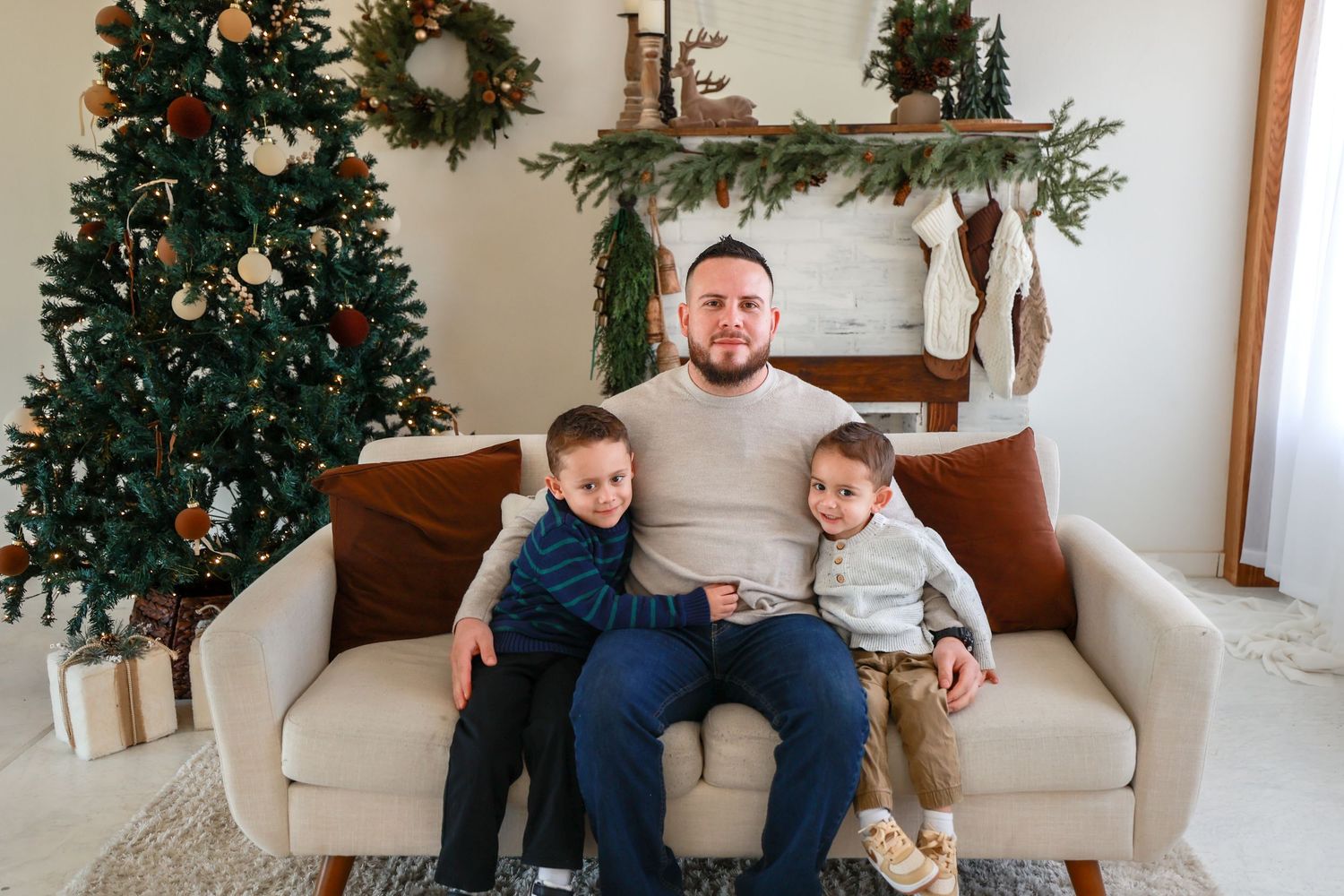 Father sits on cream sofa with two young sons beside a decorated Christmas tree and festive fireplace.