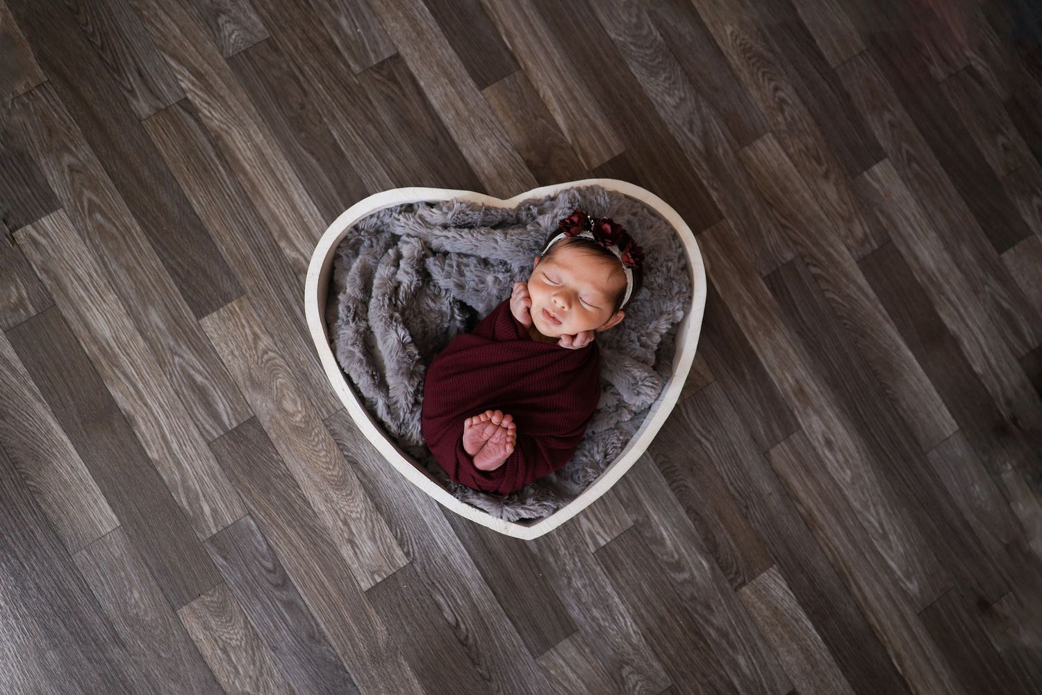 Smiling newborn baby wrapped in burgundy swaddle resting in a heart-shaped white basket.