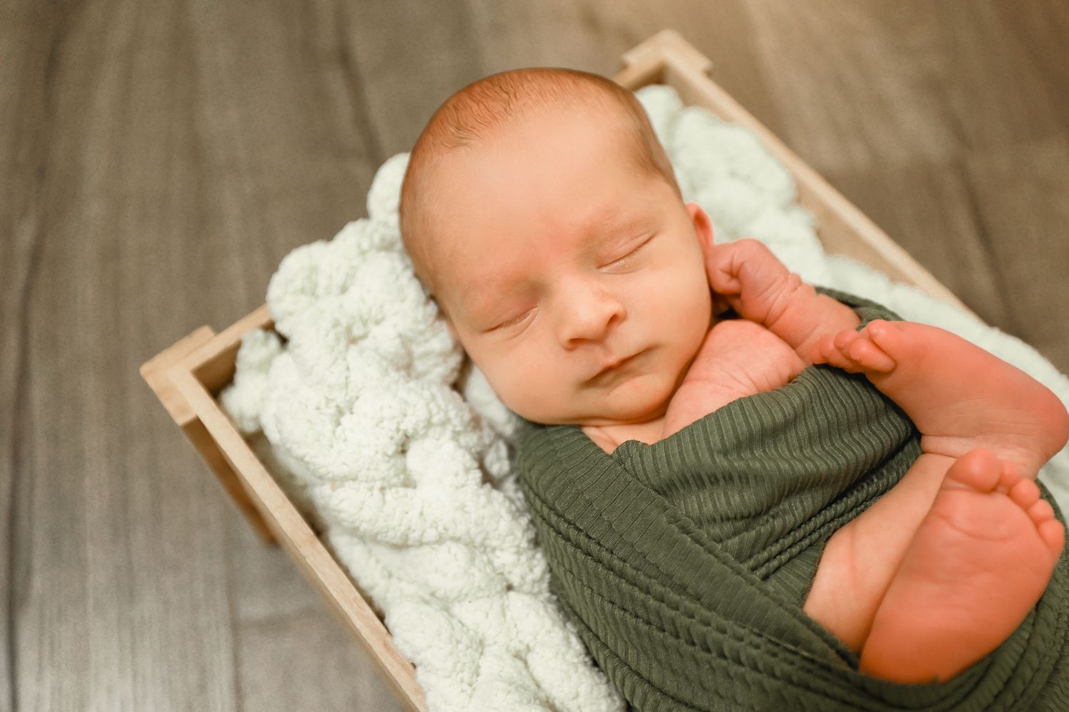 Sleeping newborn baby swaddled in green wrap resting in a rustic wooden crate.