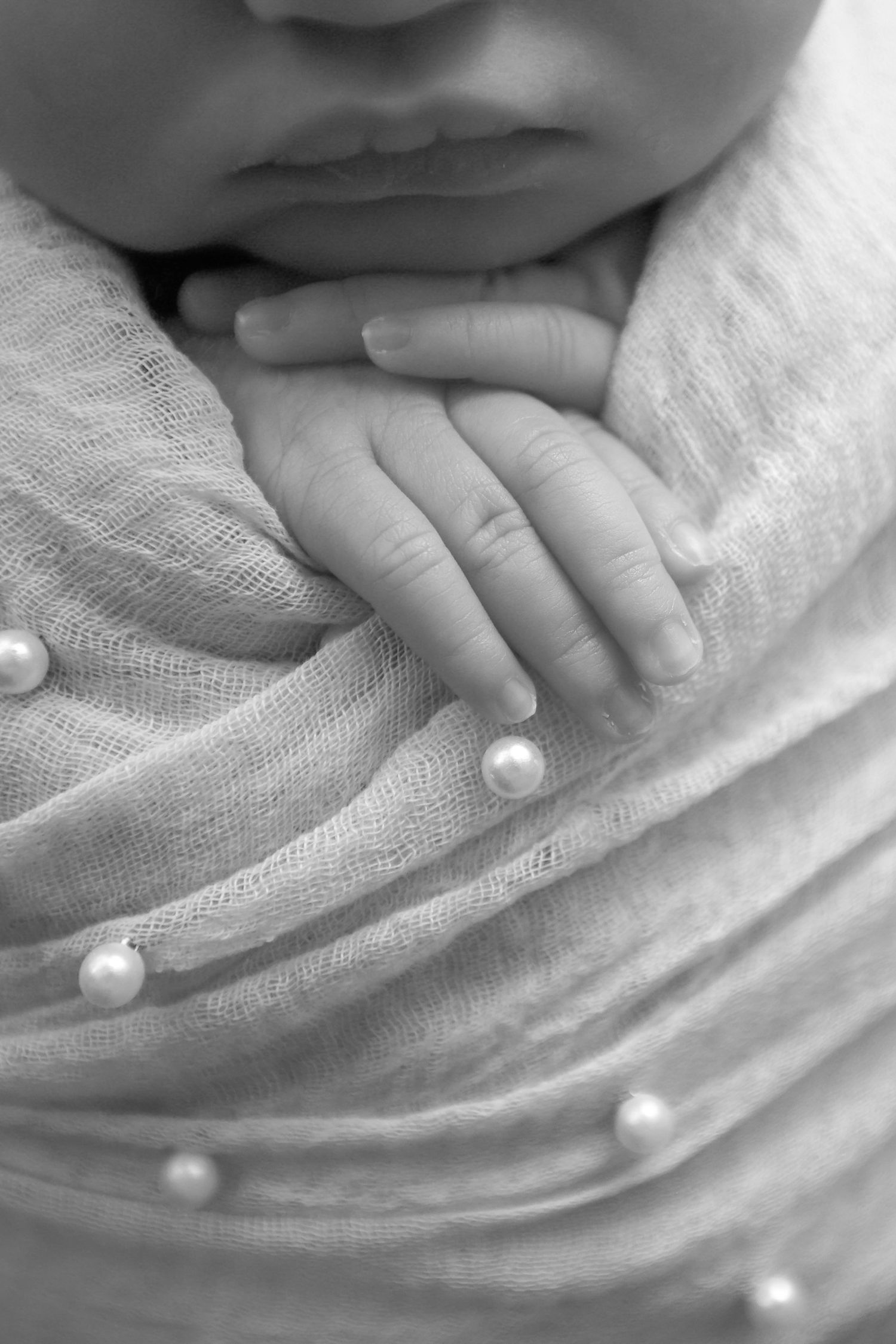 Black and white photo of a swaddled newborn baby's tiny hands resting on a soft wrap adorned with pearl beads.