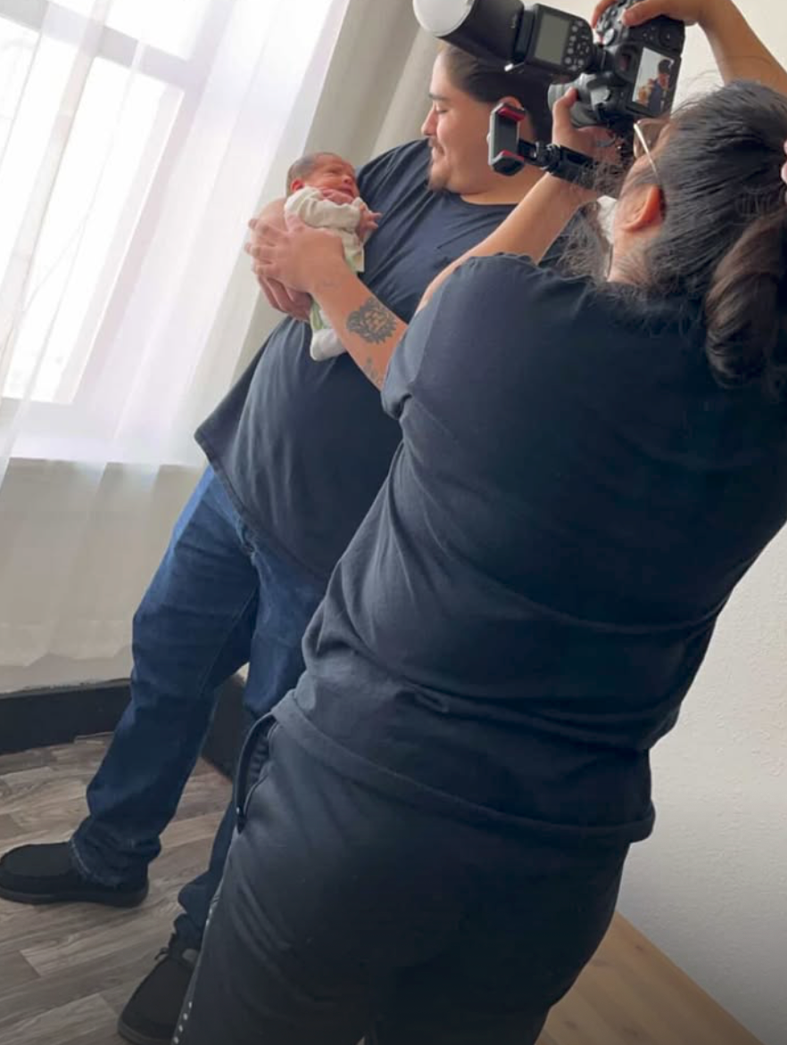 Behind-the-scenes view of professional newborn photographer shooting father holding newborn baby near window.