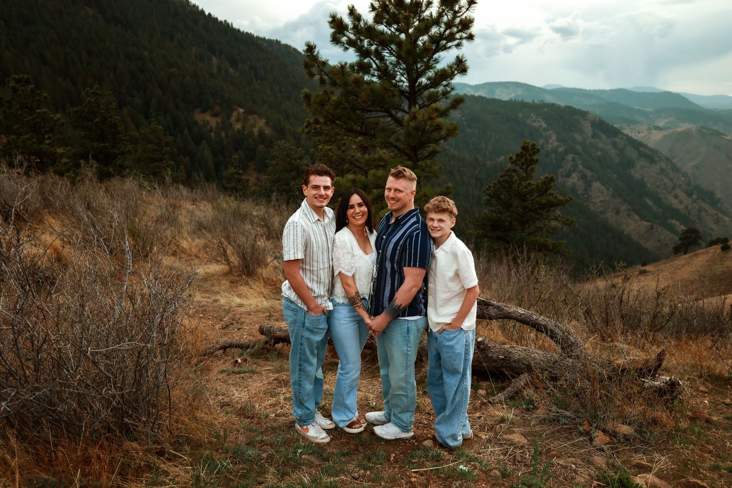 Family of four posing on a mountain trail with scenic valley and pine trees in the background.
