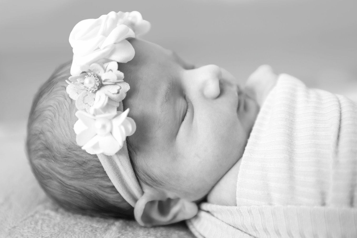 Black and white photo of a sleeping newborn baby girl wearing a delicate white floral headband, swaddled in white.
