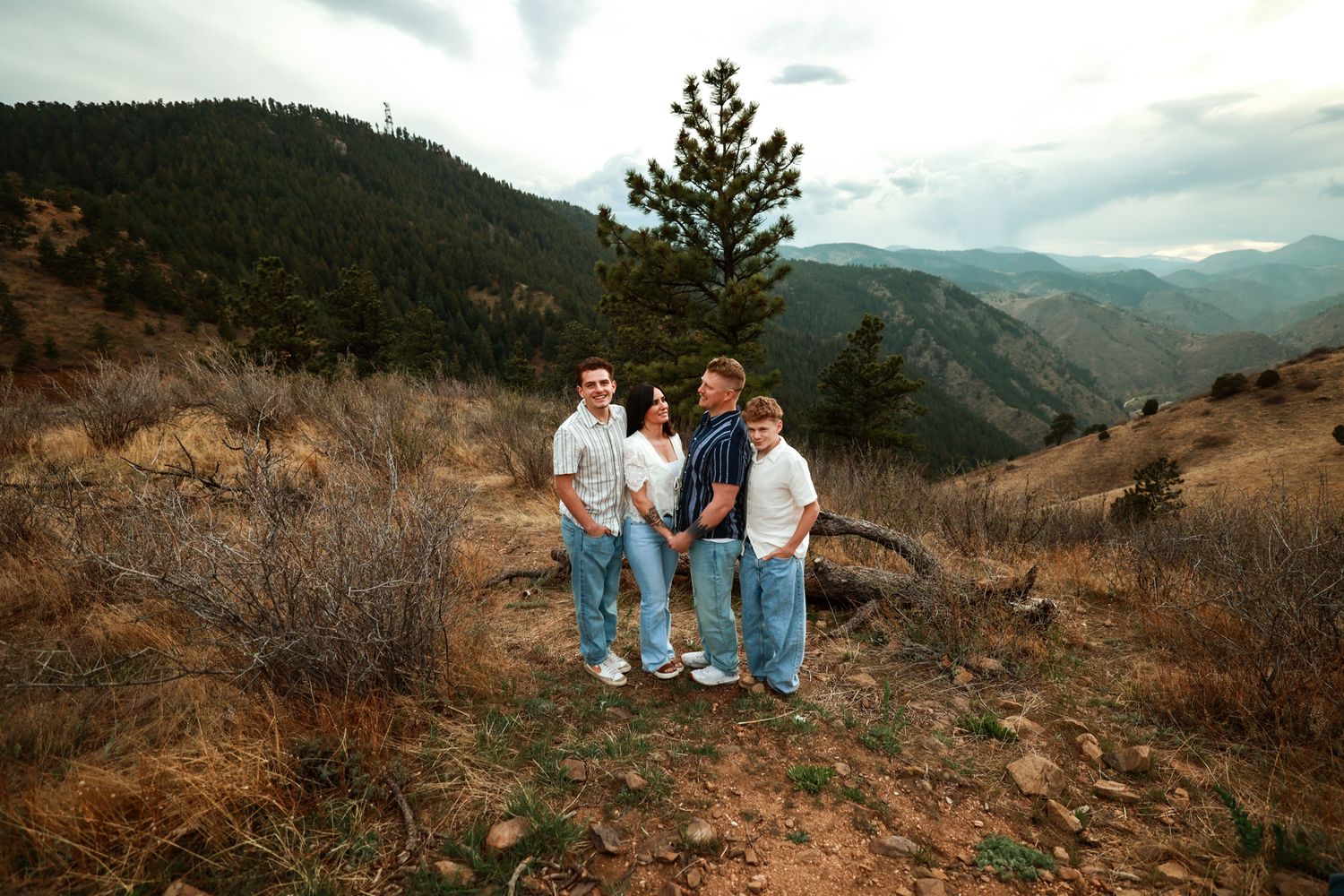 Family of four posing on a mountain trail with scenic valley and pine trees in the background.