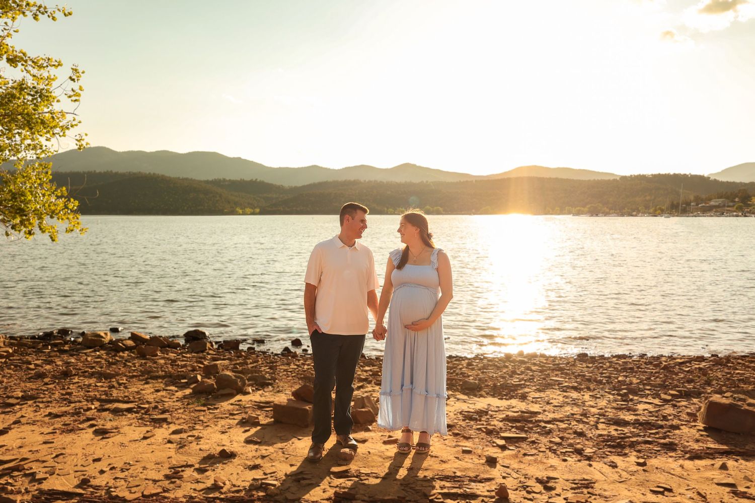 Expecting couple holding hands at golden hour sunset by a scenic mountain lake.