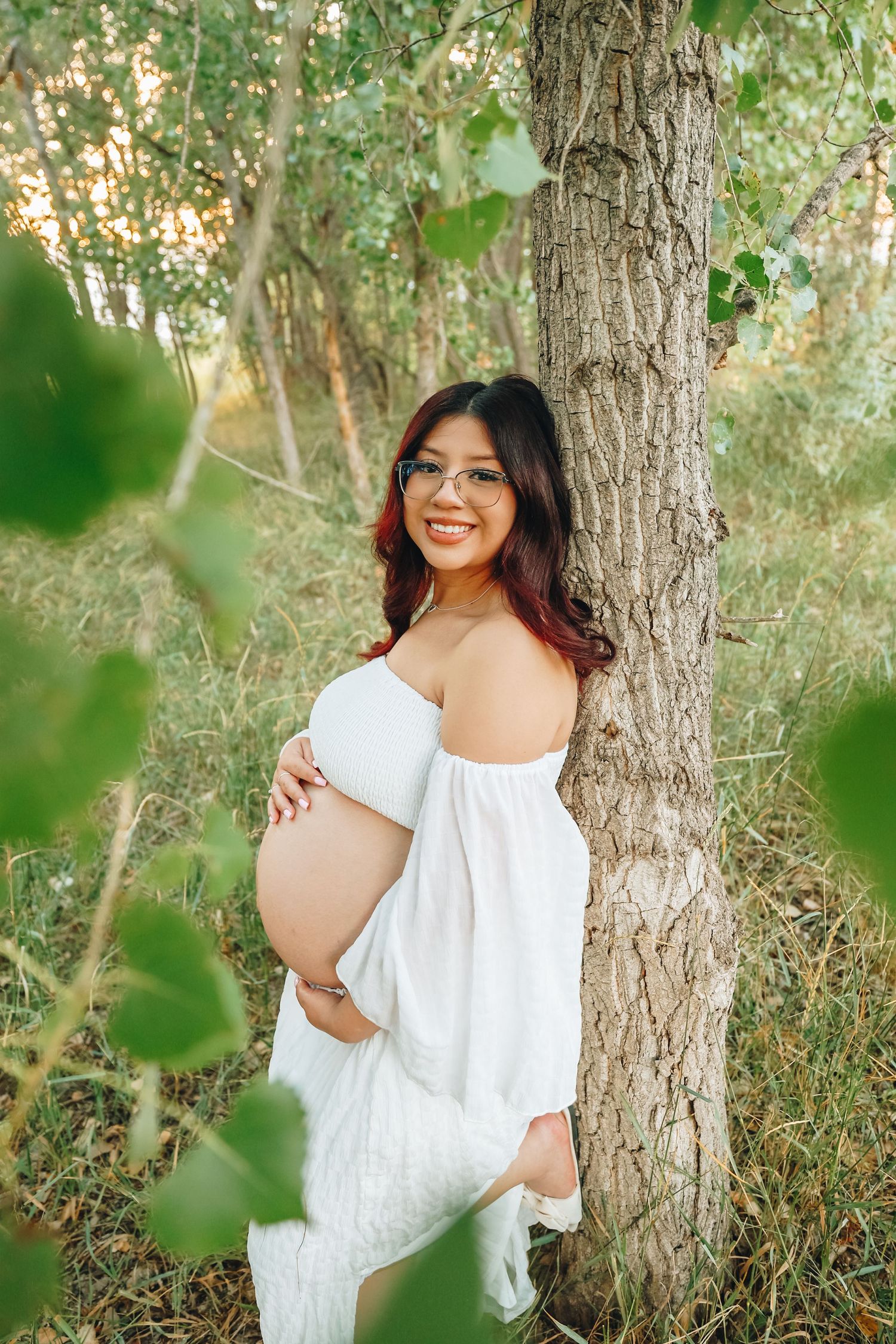 Smiling pregnant woman in white off-shoulder dress leaning against tree in lush green forest.
